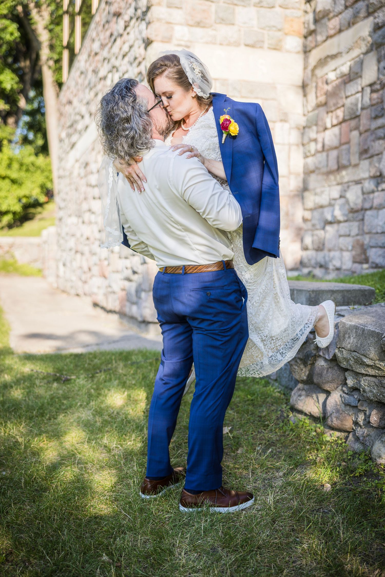 Romantic moment captured between couple in blue suit and white dress by stone wall.