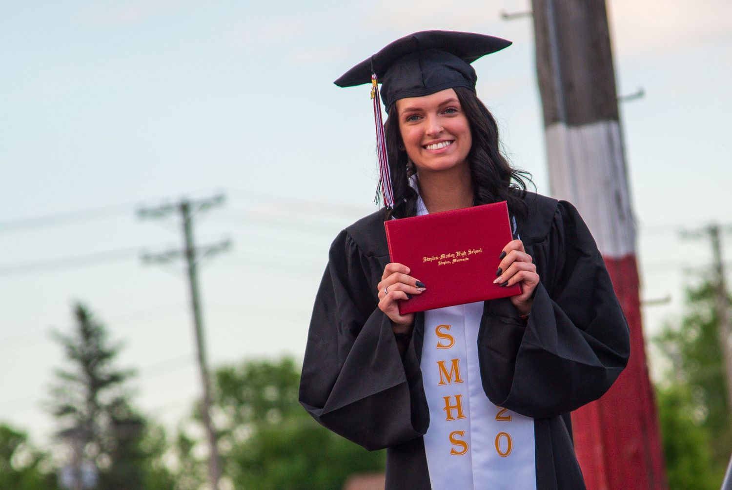 Graduate in cap and gown holding red diploma during outdoor ceremony.