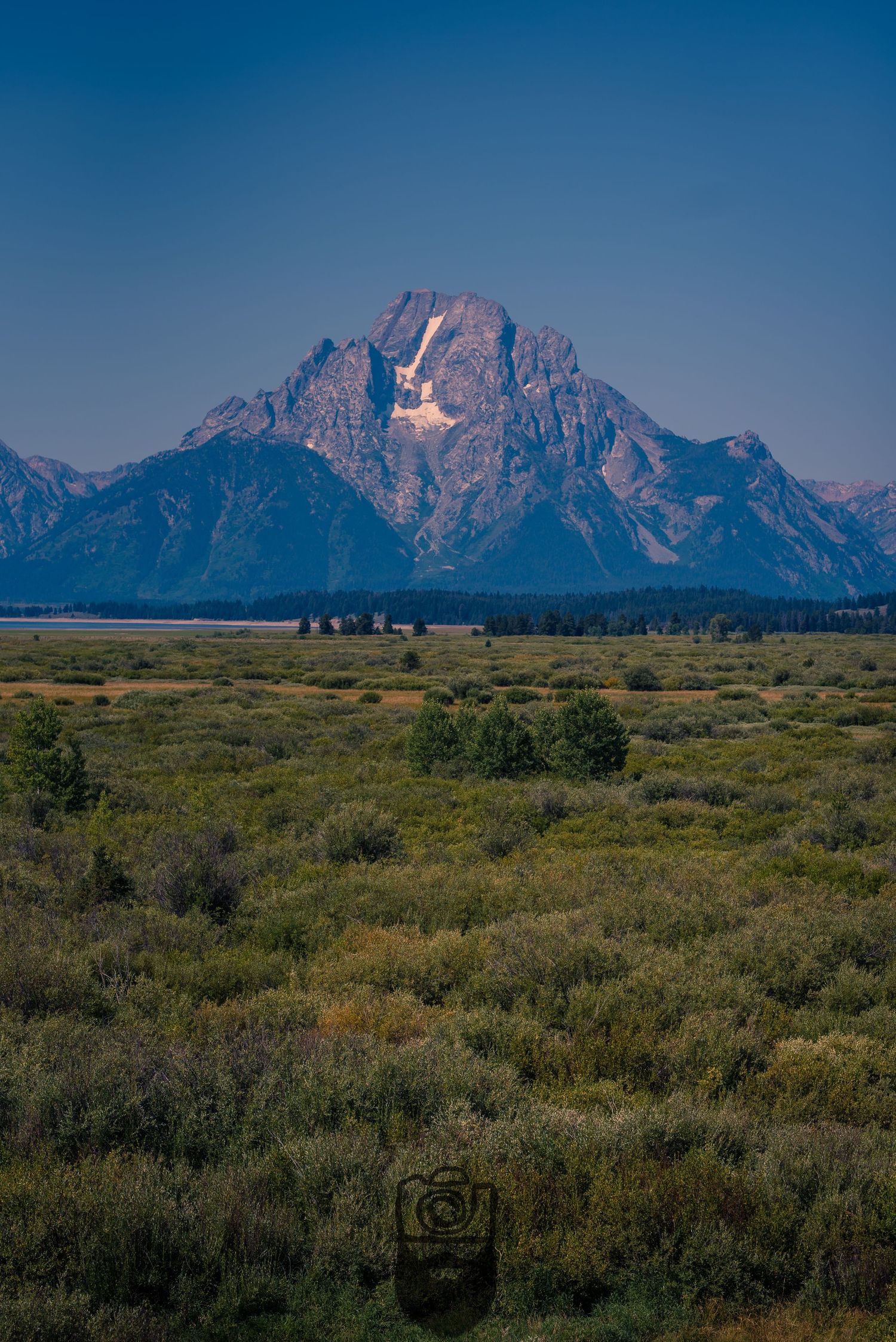 Grand Teton mountain range rises above sage brush plains under clear blue sky.