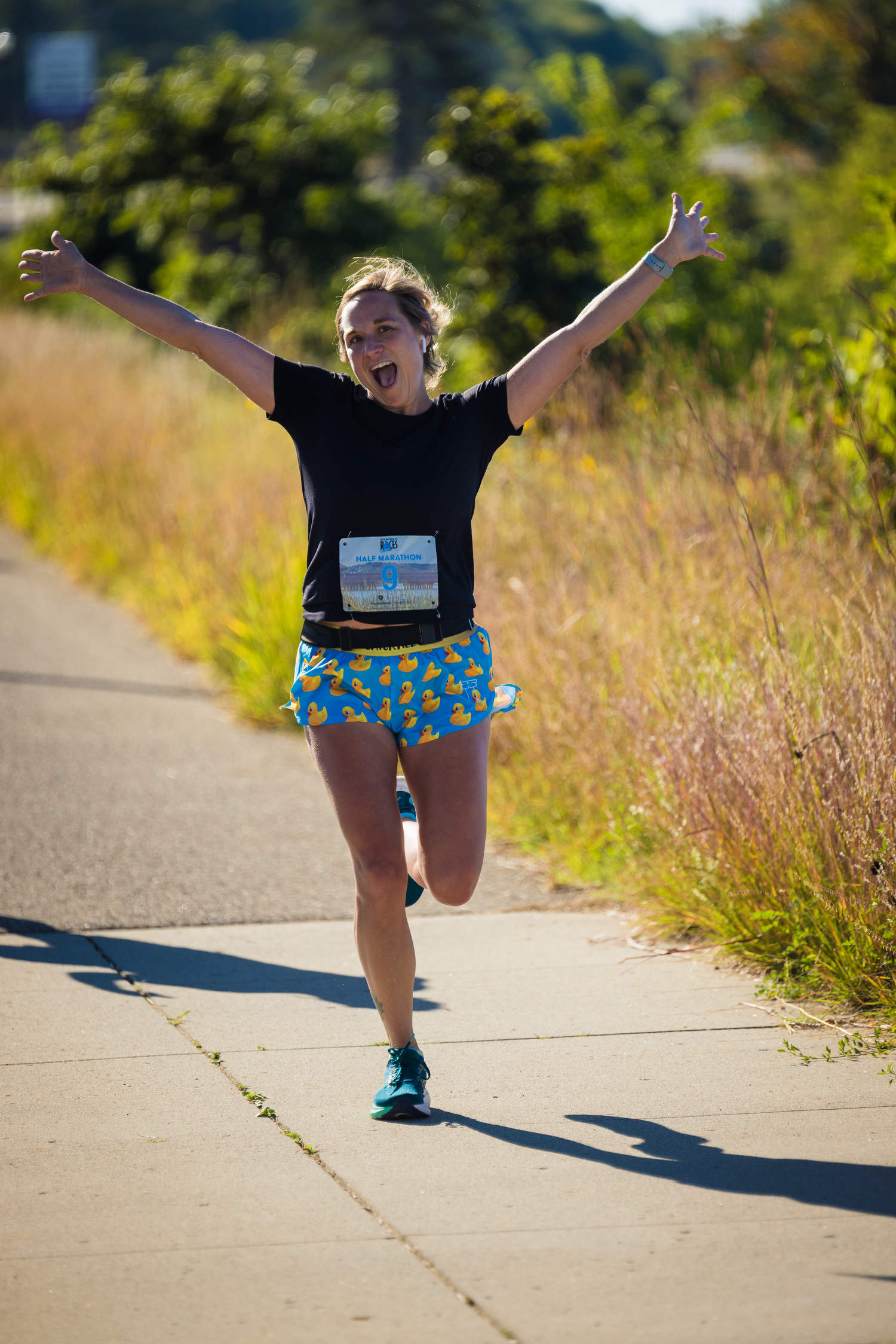 Close-up of running shoes and legs on a paved path with tall grass on both sides.