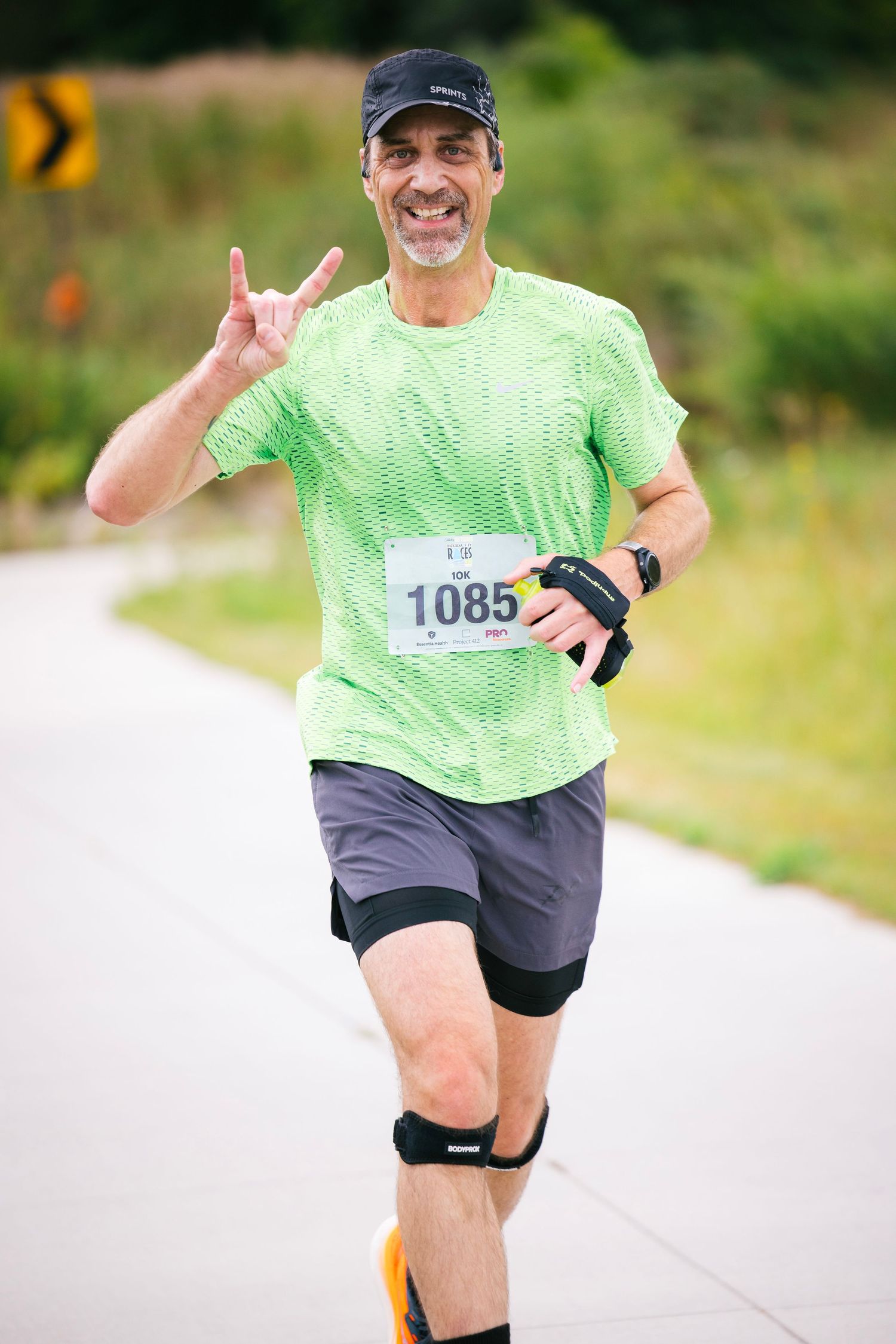 Runner in bright green shirt and black shorts smiling while jogging on a paved path during a race event.