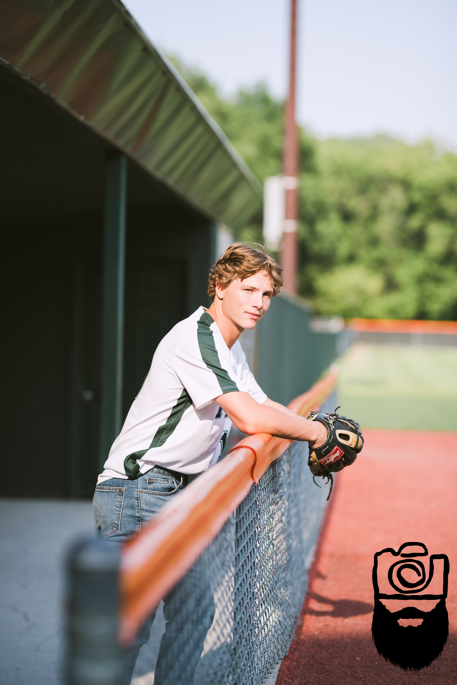 Person leaning against baseball dugout fence in white uniform during sunny day.
