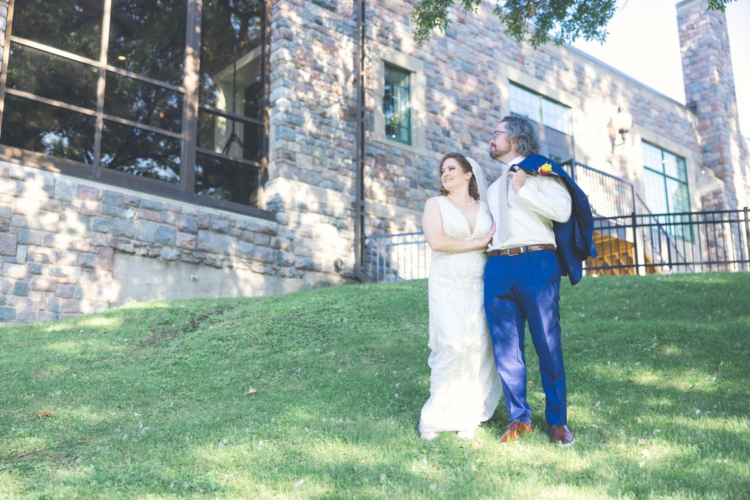 Couple embracing outdoors near a stone wall and grassy hill.