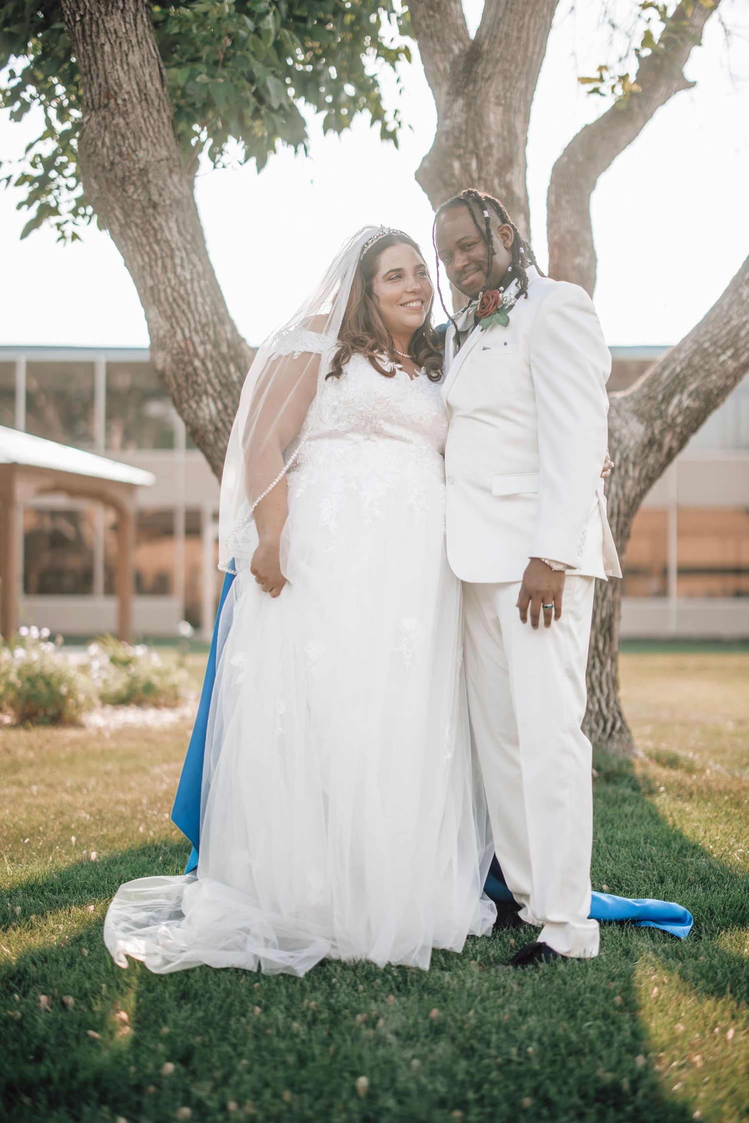 A couple poses for romantic photos on grass under large trees in white formal attire.