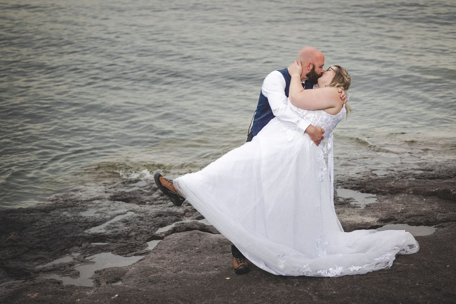 A couple in wedding attire sits together on rocks by the water.