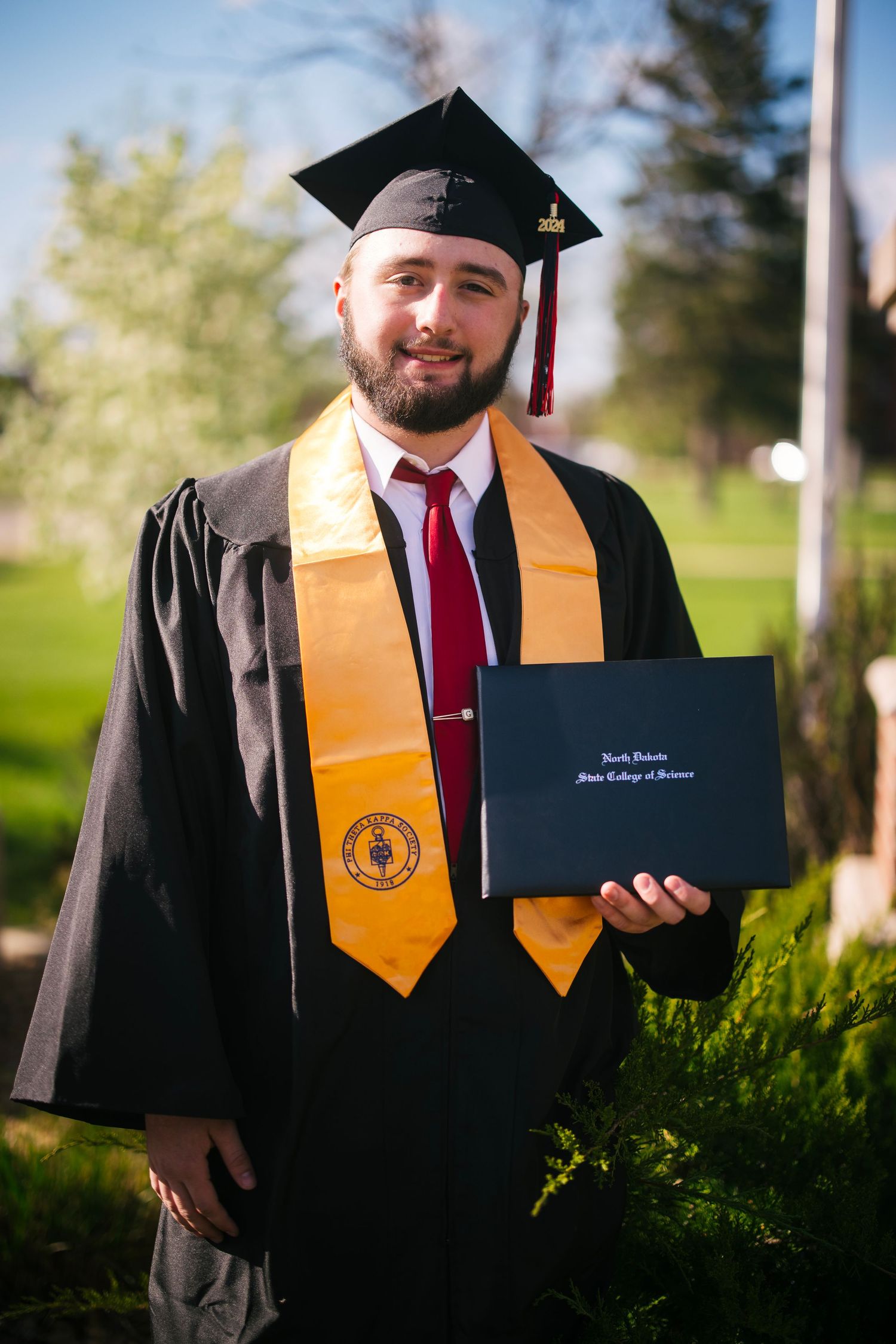 Graduate in black cap and gown with gold honors stole holding diploma during outdoor ceremony.