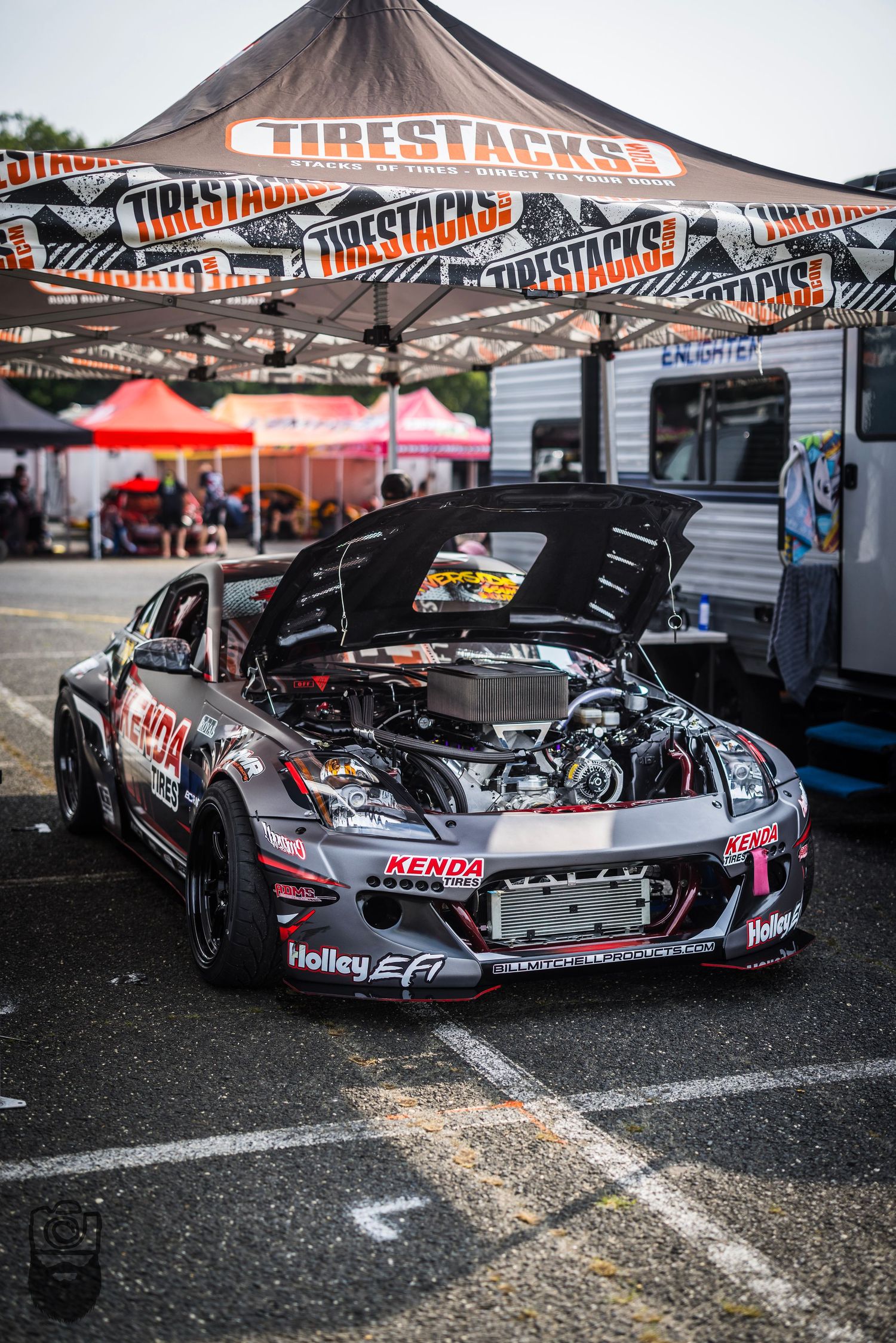 Race cars parked under Tirestacks vendor tents at motorsport event.
