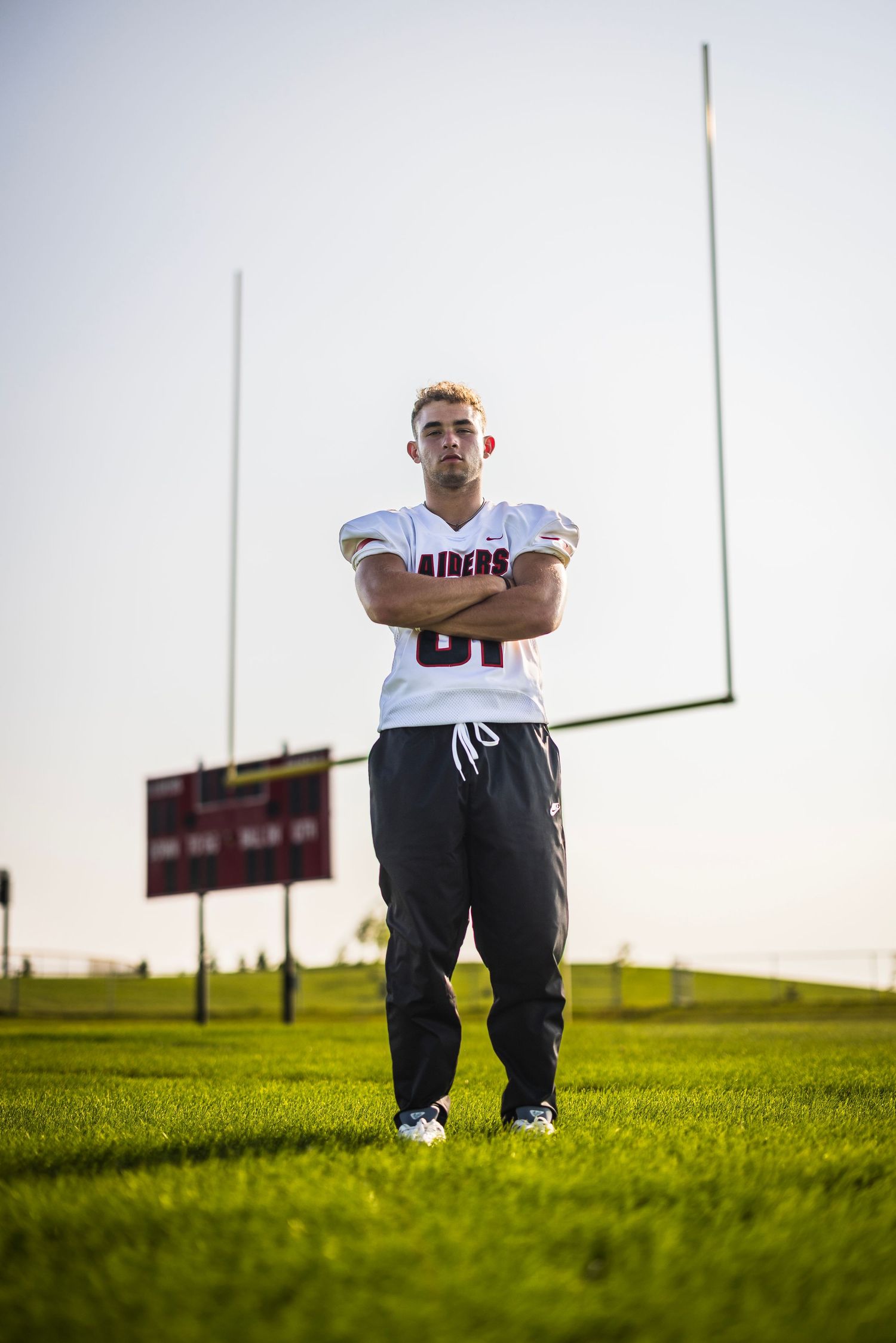 Athlete in white uniform sitting on football field at sunset with goal posts in background.