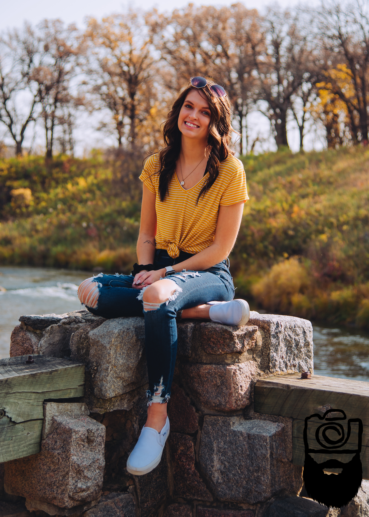 Person in yellow top and jeans sitting on stone wall by river during autumn.