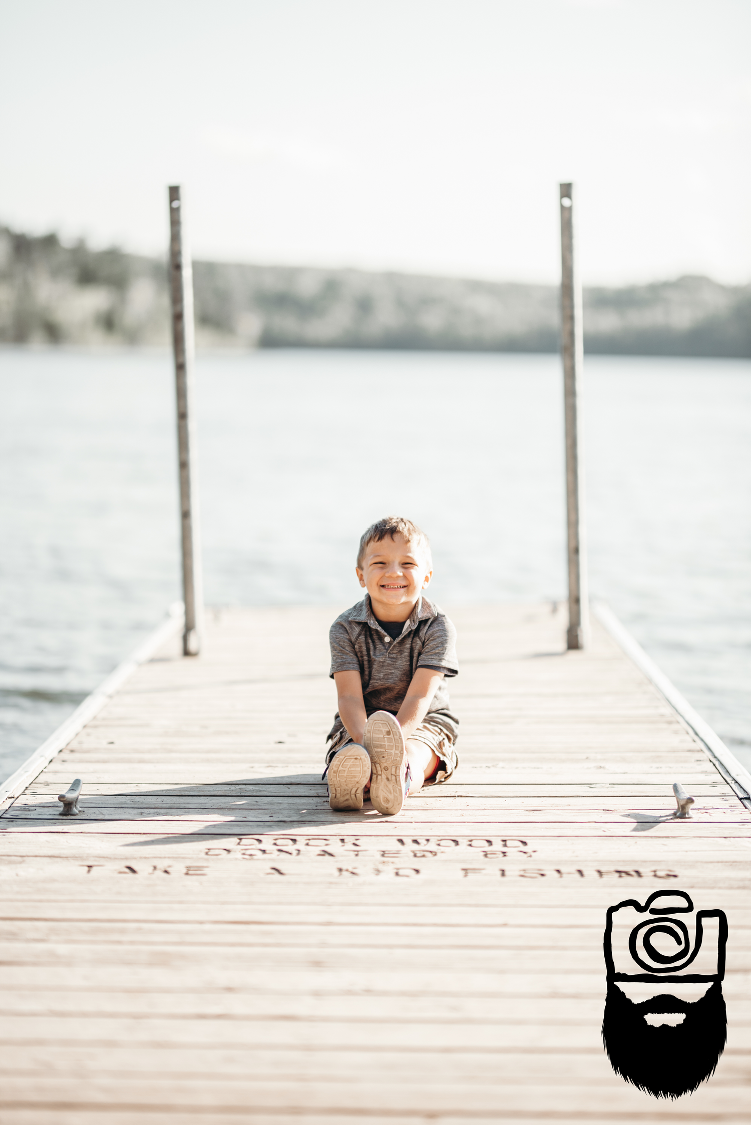 Child sitting on wooden dock by tranquil lake on sunny day.