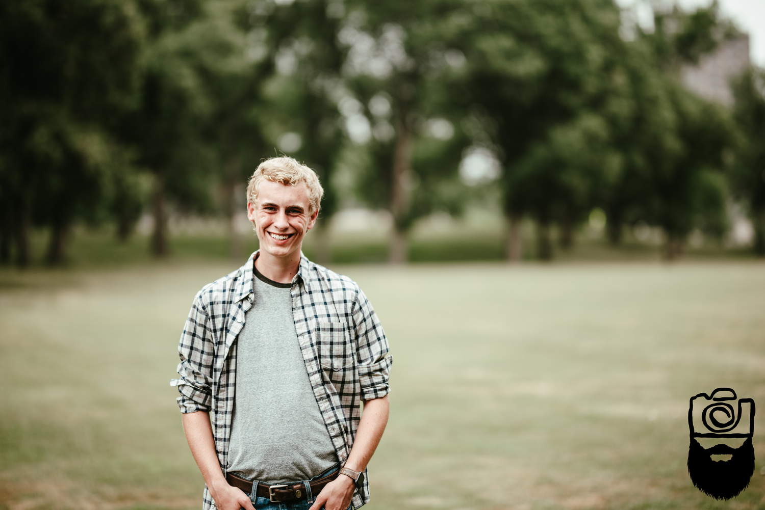 A man in a plaid shirt stands casually in a green outdoor setting.