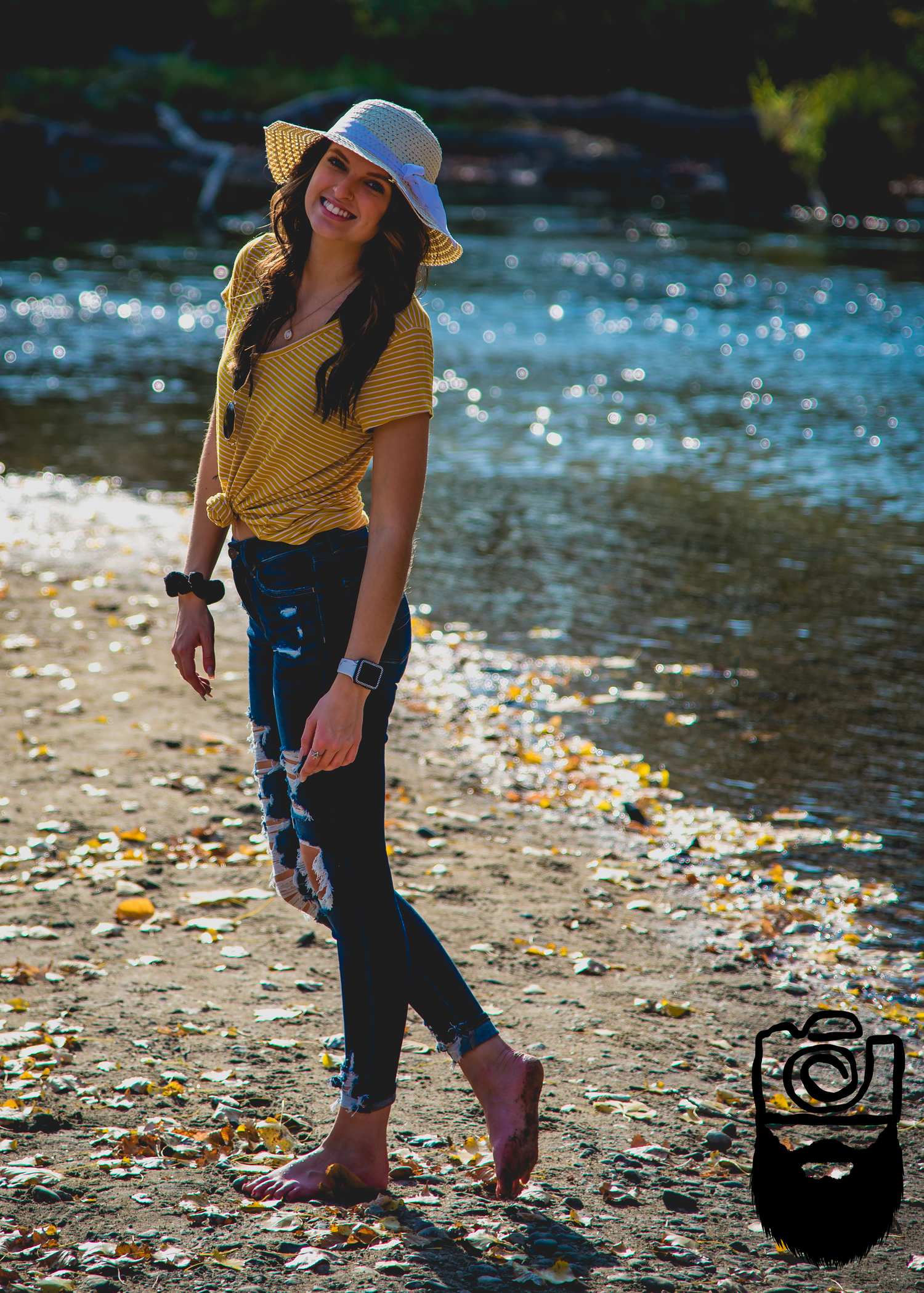A person in a yellow shirt and jeans walks along a riverbank covered in autumn leaves.