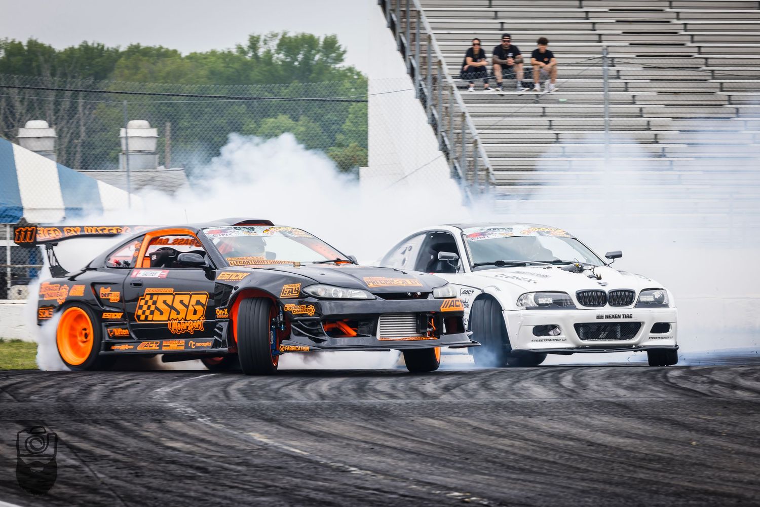 Professional drift cars creating smoke clouds during tandem battle on race track.