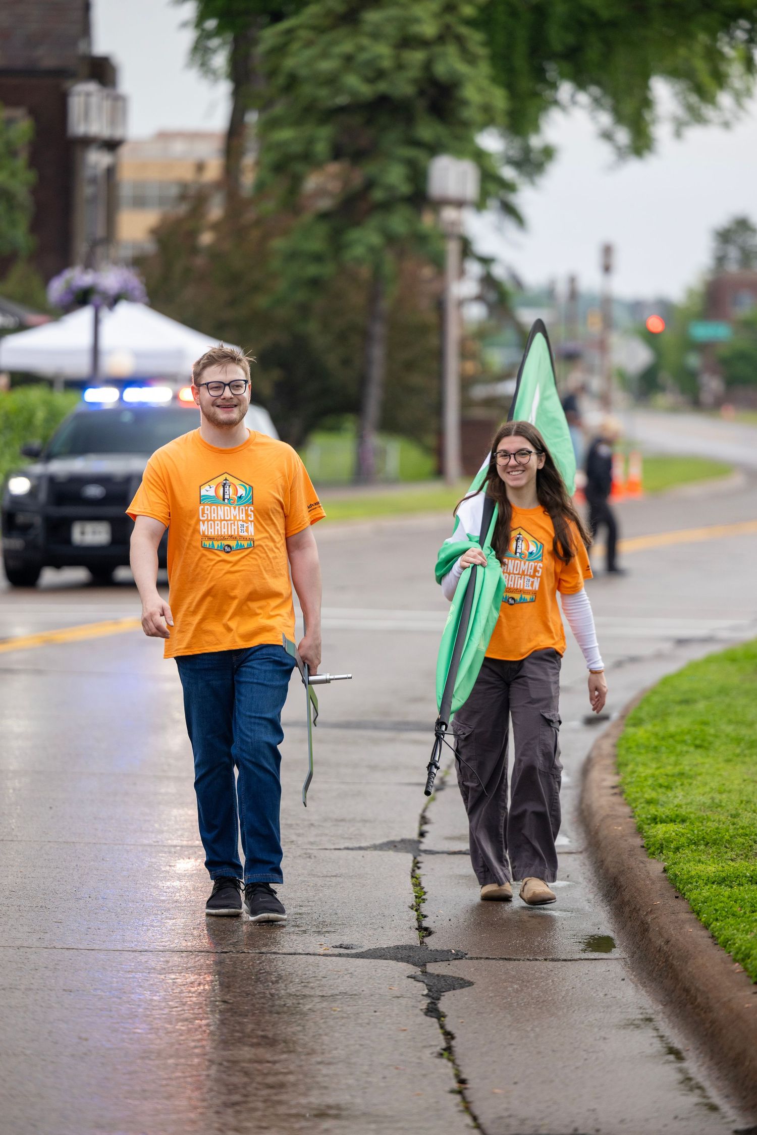 Volunteers in orange shirts carrying green flags walk along a wet sidewalk.