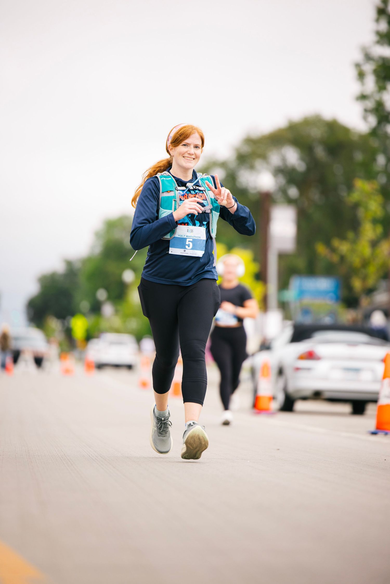 Runner in blue jacket leaps energetically during outdoor race with parked cars in background.