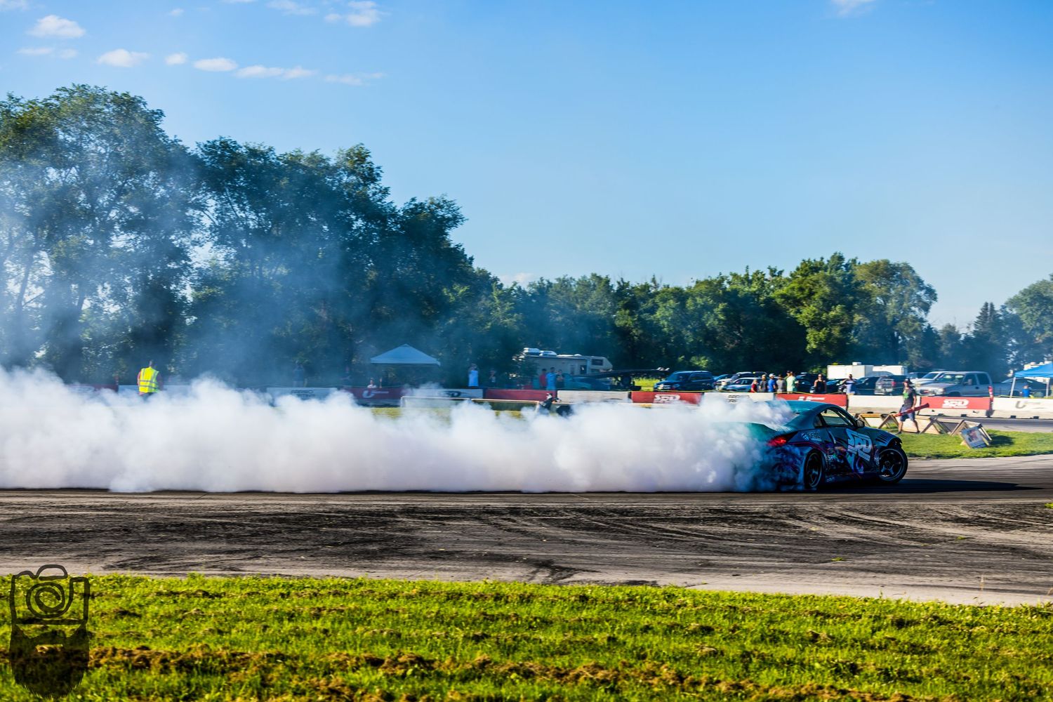 Cars perform drift maneuvers creating smoke trails across sunny racetrack.
