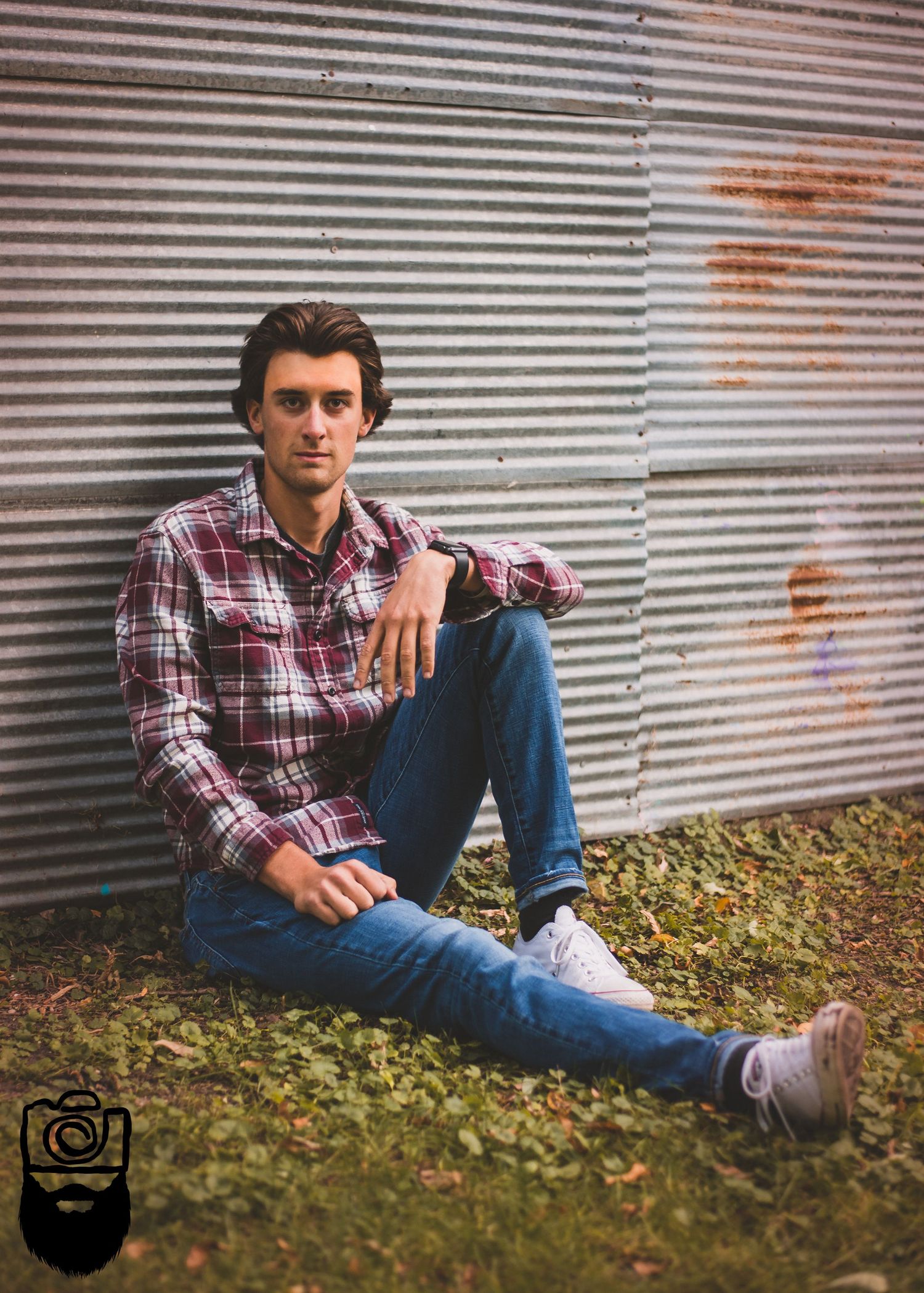 Person in plaid shirt and jeans relaxing against corrugated metal wall outdoors.