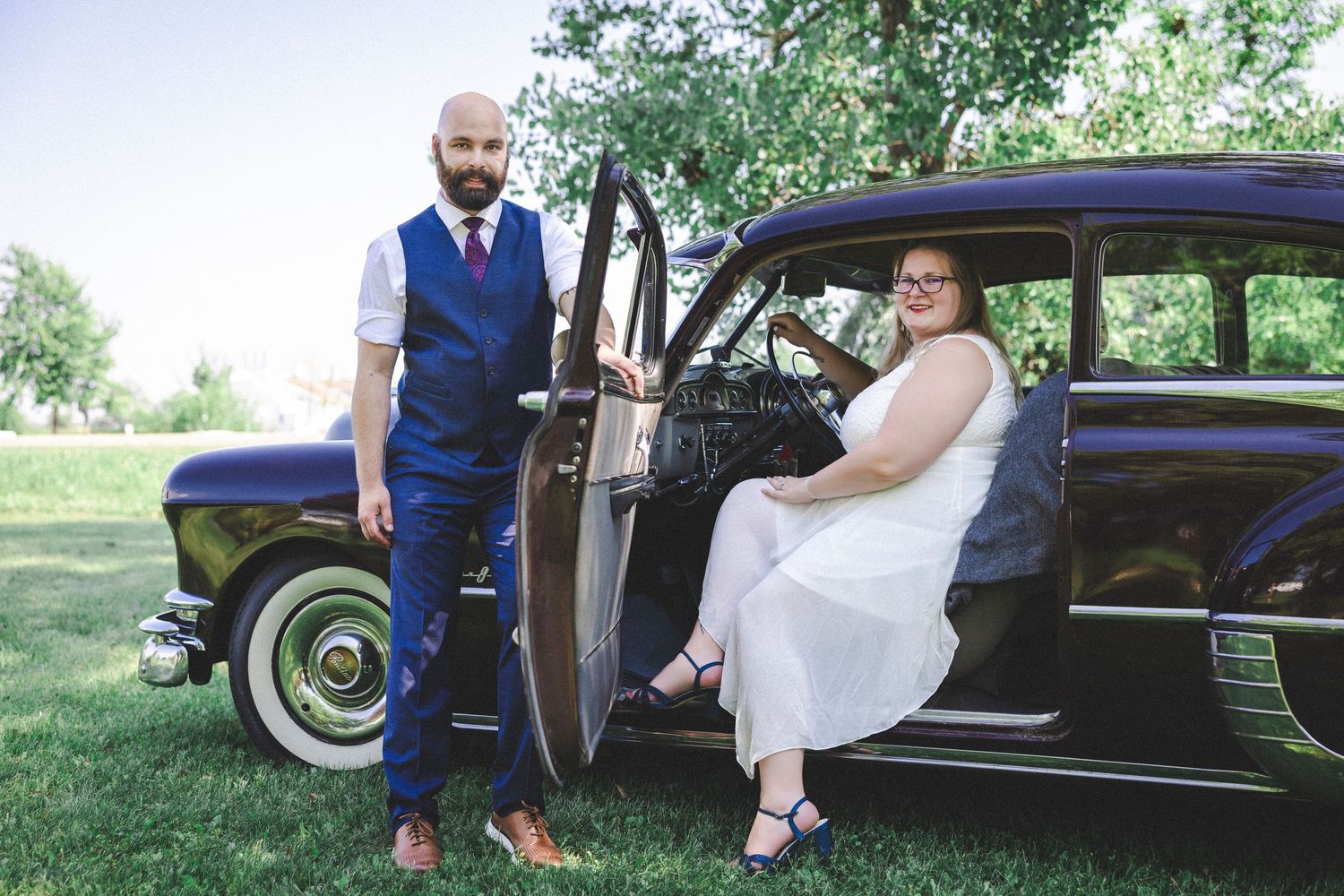 Wedding portraits taken with a vintage black car in a rustic outdoor setting.