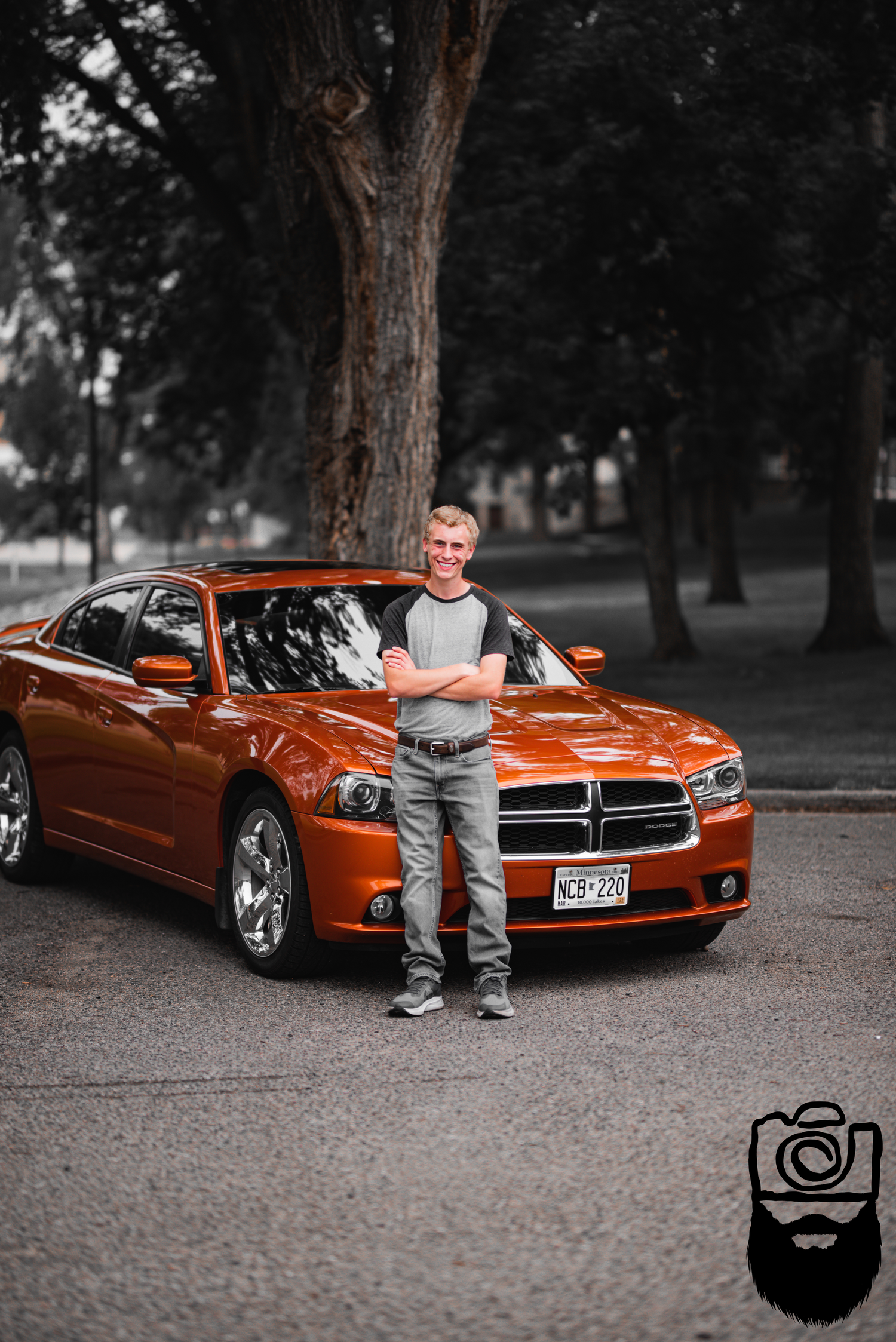 A bright orange Dodge Charger parked under trees with someone posing beside it.