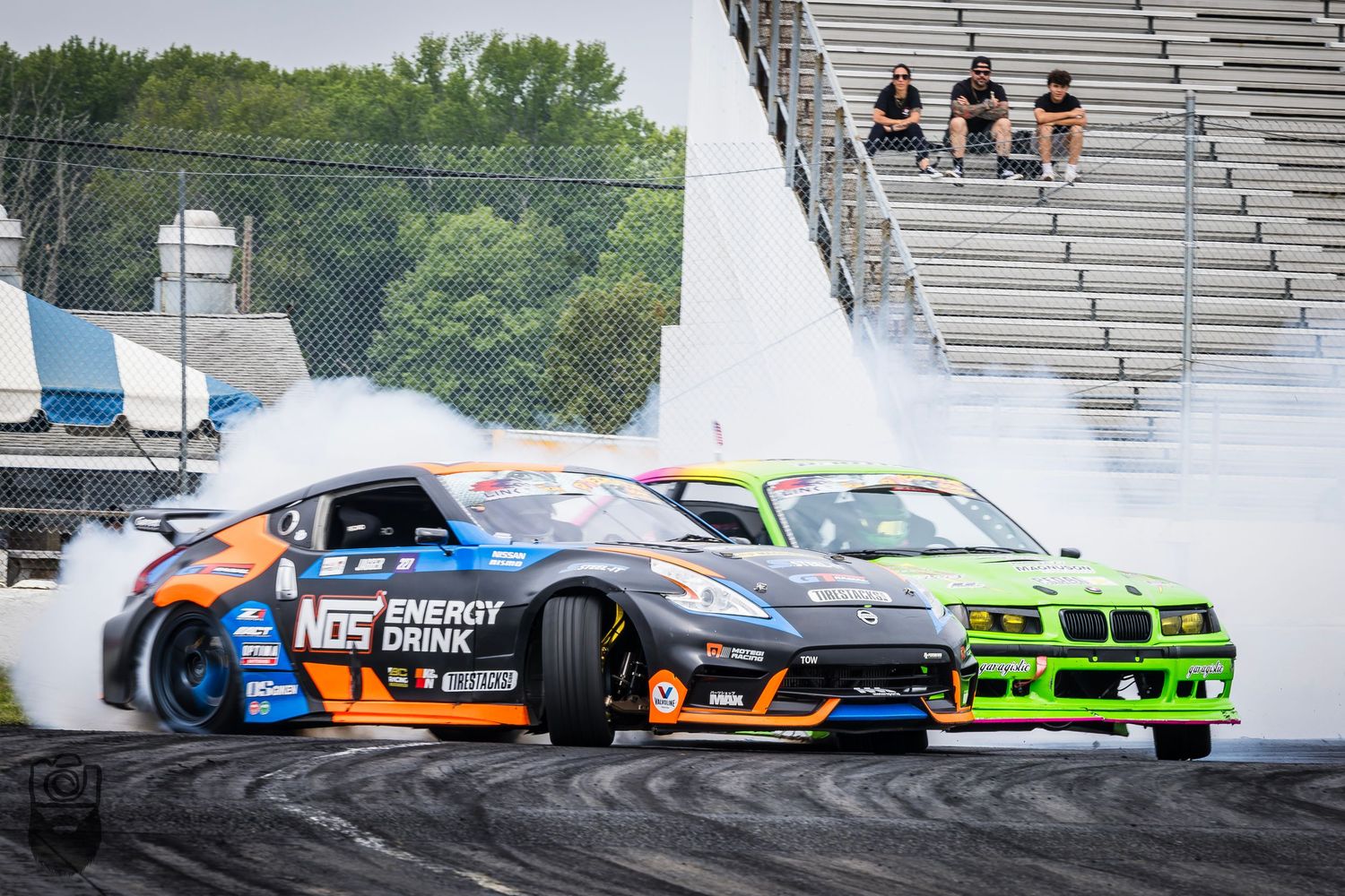 Multiple drift cars creating tire smoke while competing on track with spectator stands.