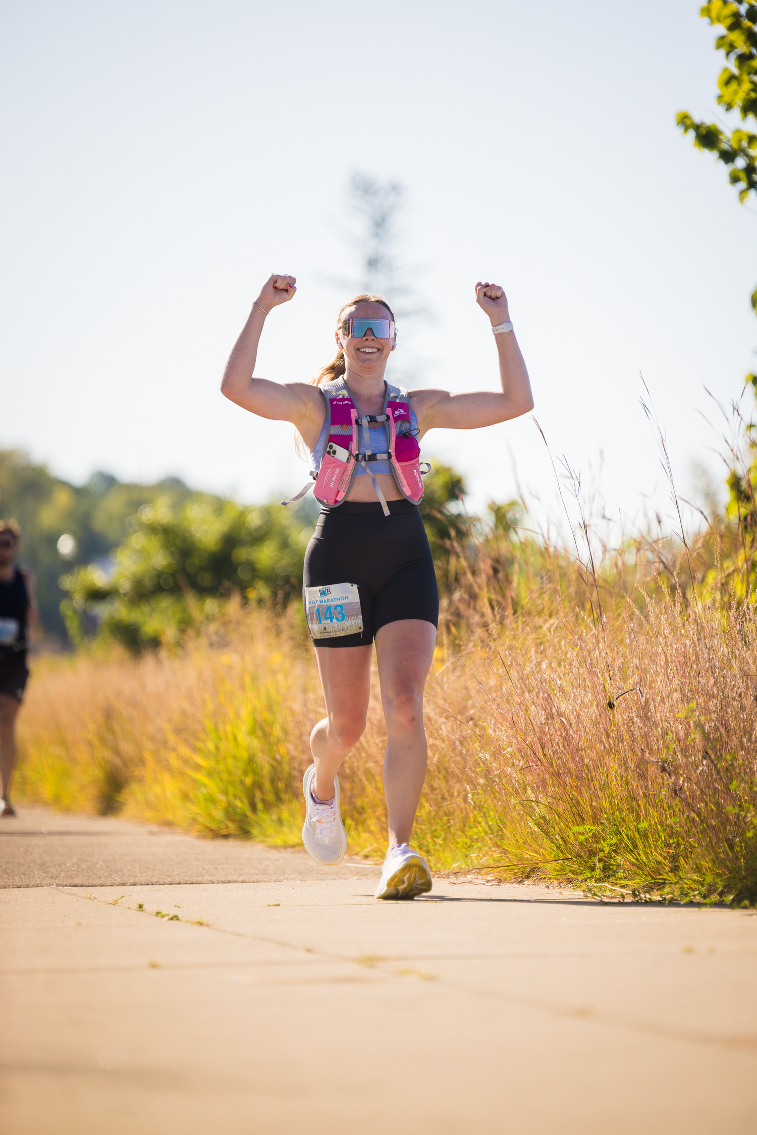 Runner celebrates with raised arms while jogging on a sunlit trail during race competition.