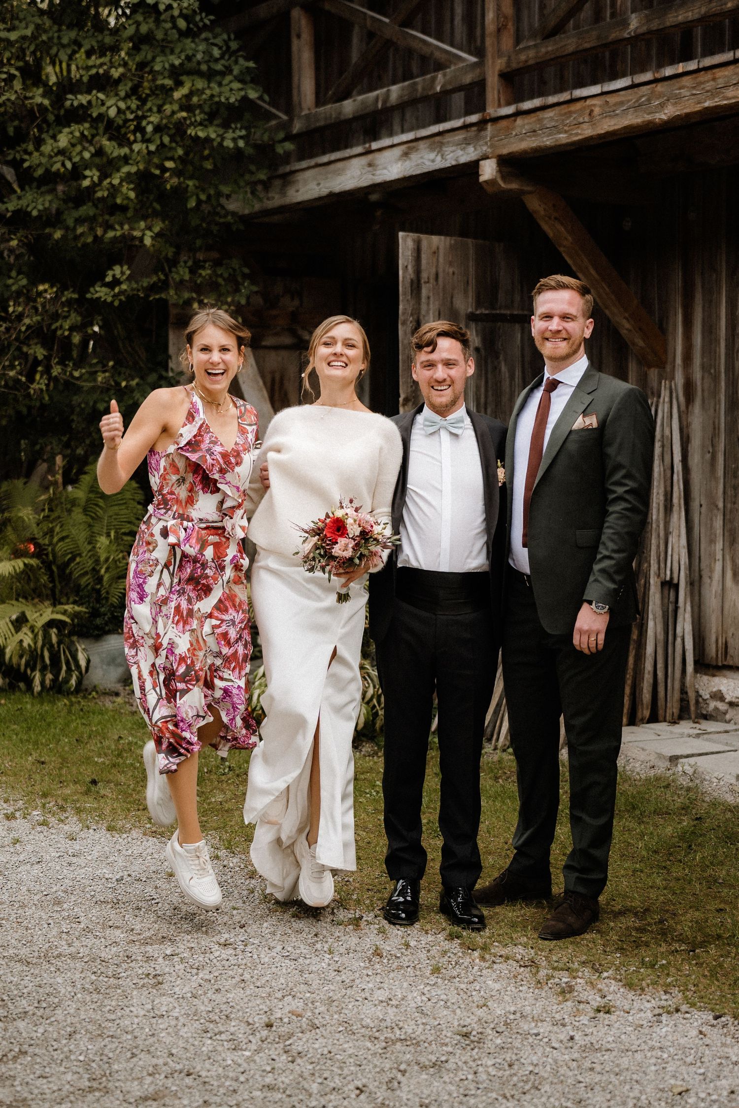 Wedding party in mix of formal attire and summer dresses poses casually by wooden barn.