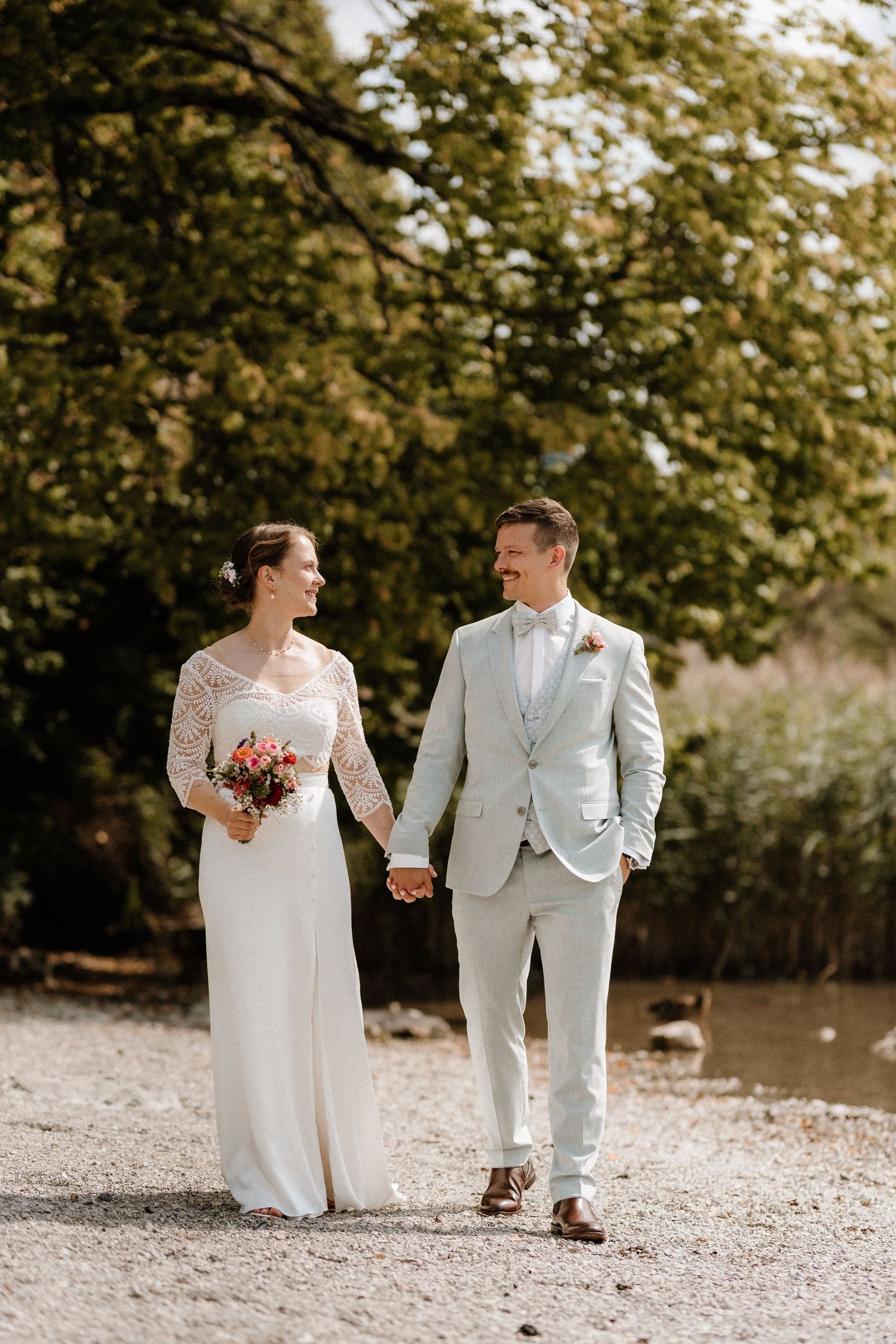 A wedding couple walks hand in hand along a lakeside path surrounded by nature.