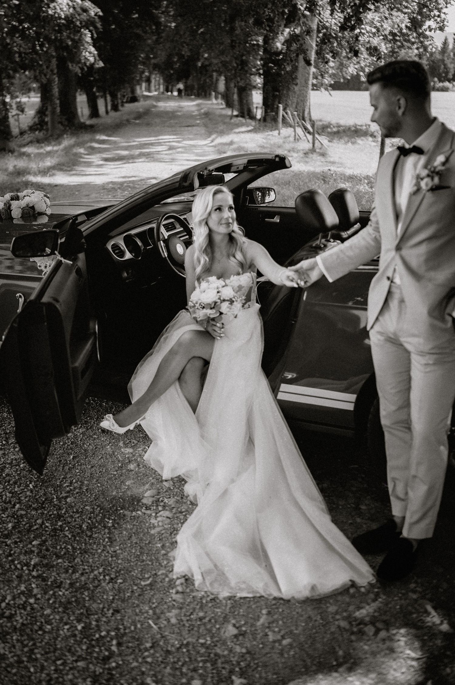 Black and white photo of a couple's romantic moment by a vintage car.