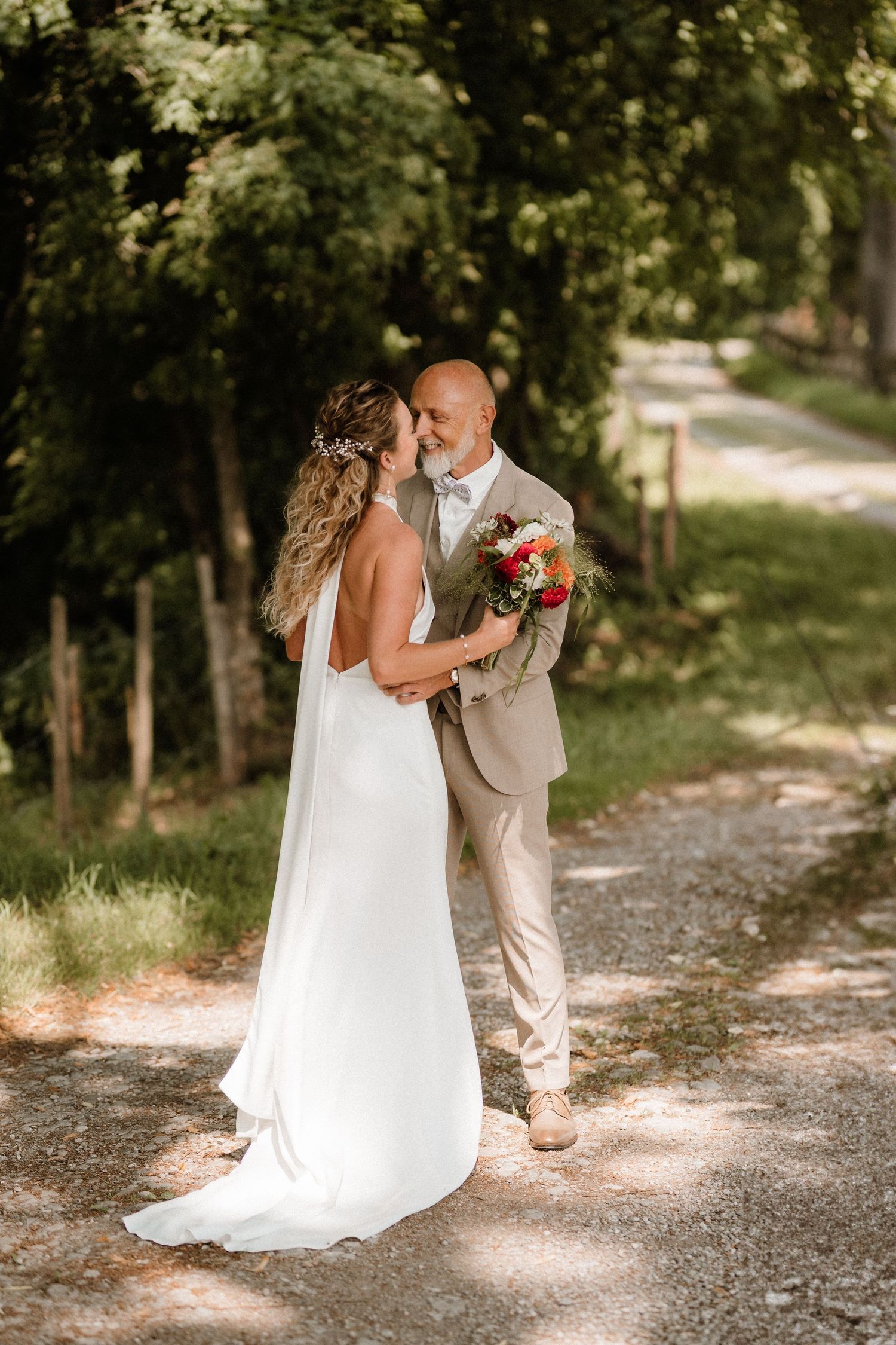 Wedding couple embraces on a rustic gravel path surrounded by lush summer trees.