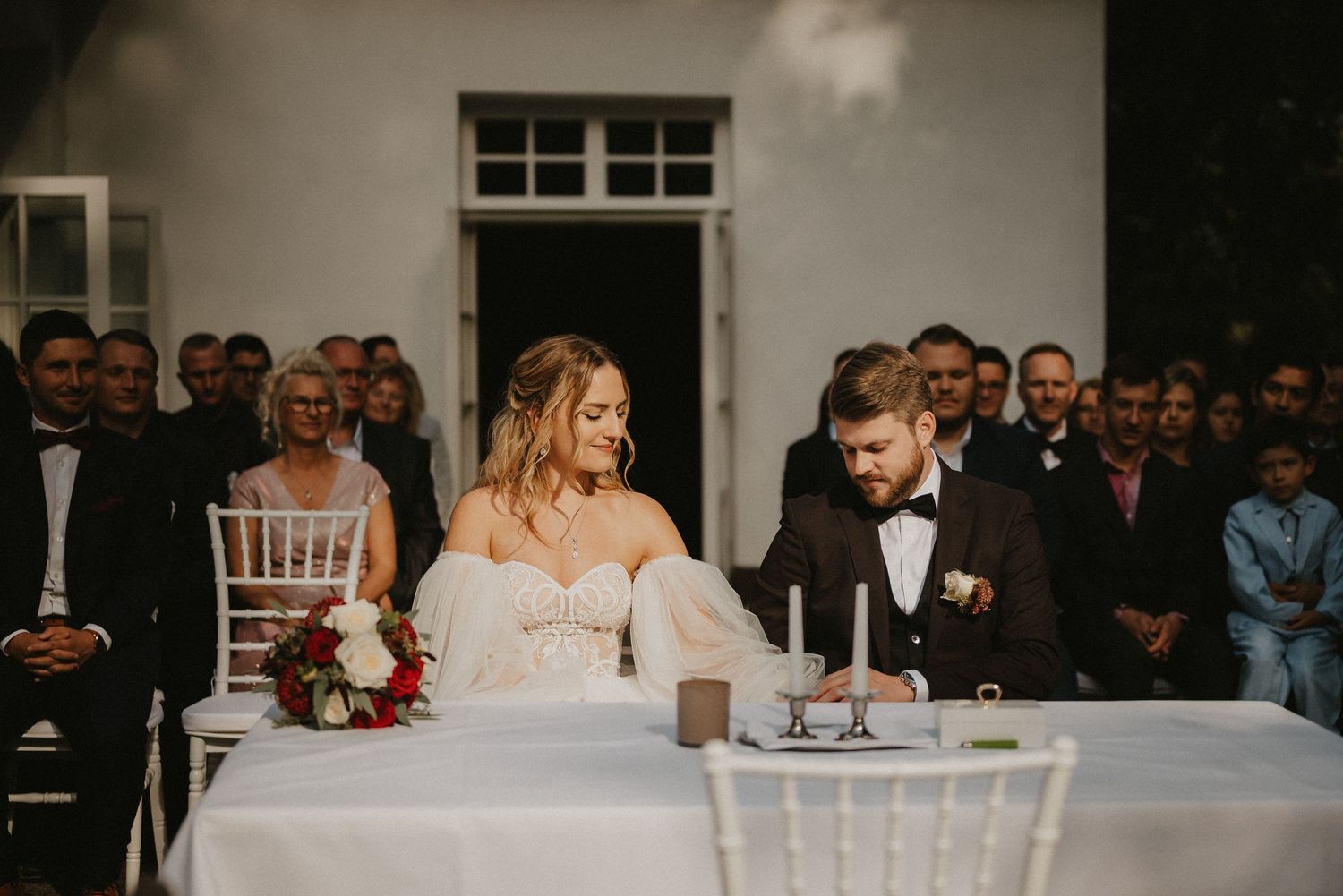 Elegant outdoor wedding ceremony with guests seated in white chairs against a white building backdrop.