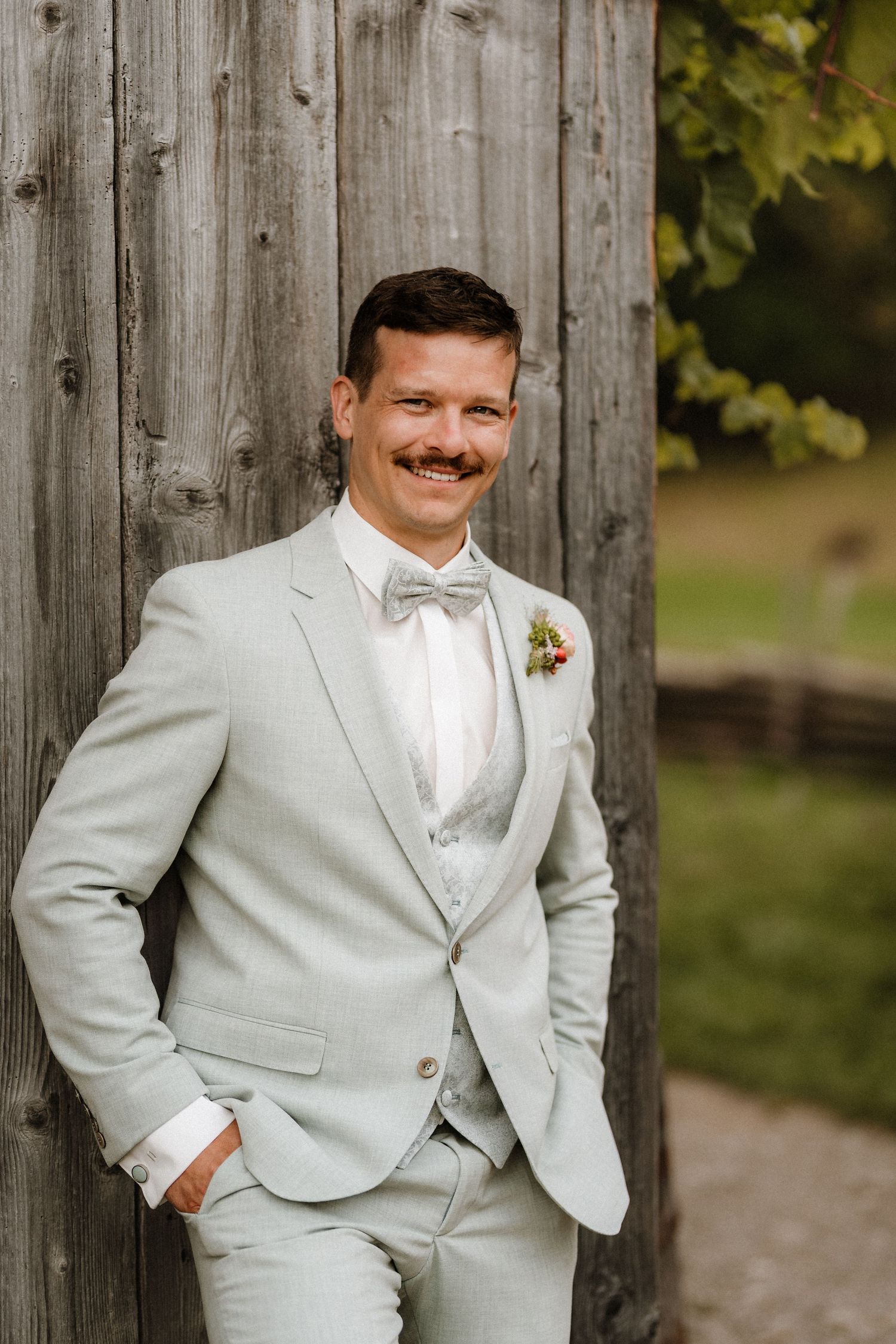 Groom in light gray suit poses confidently against rustic wooden wall.