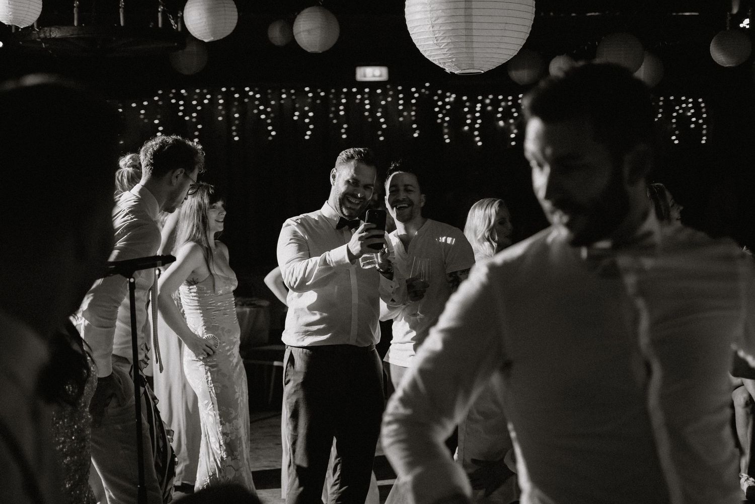 Black and white photo of guests dancing at a reception with paper lanterns and string lights.