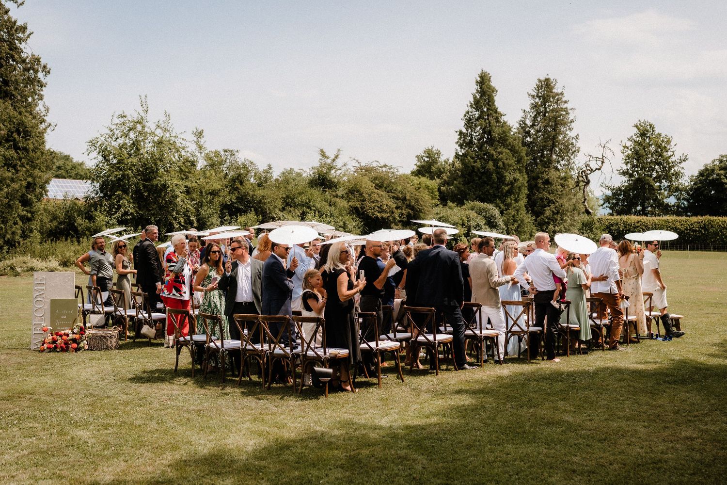 Long rows of wedding guests seated outdoors under a sunny sky with trees in the background.