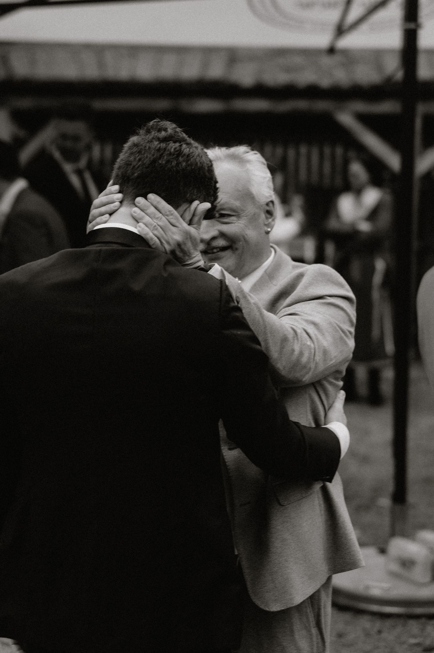 Two people share an emotional embrace at an outdoor gathering, captured in black and white photography.