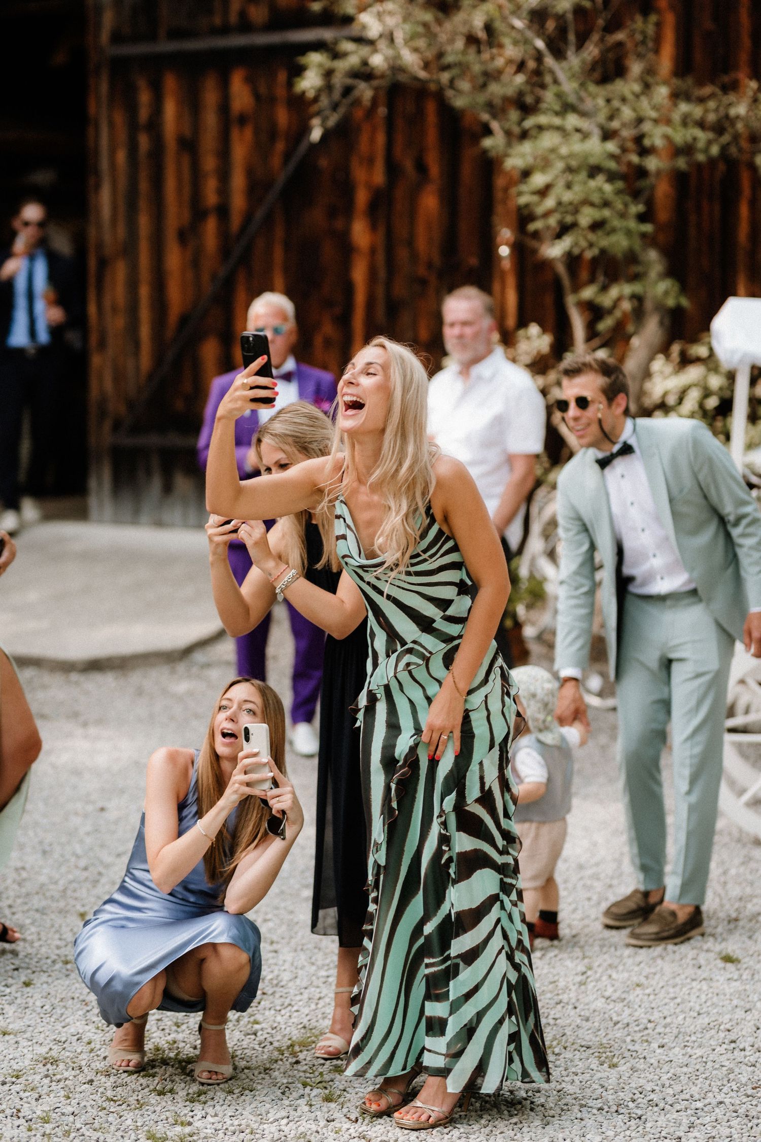 Wedding guests laughing and taking photos outside a rustic wooden barn.