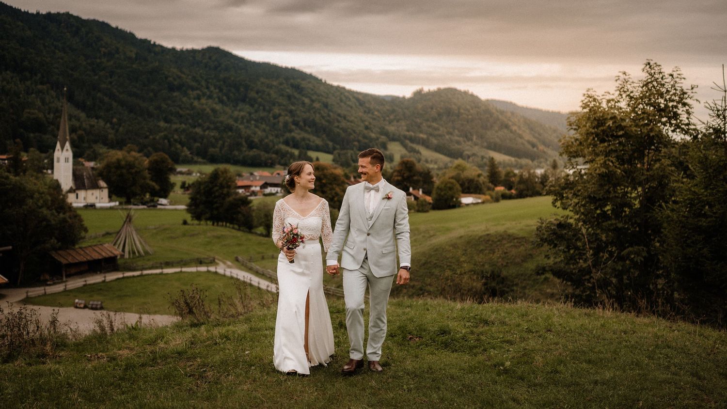 Wedding couple walking hand in hand through mountain meadow at dusk with church in background.