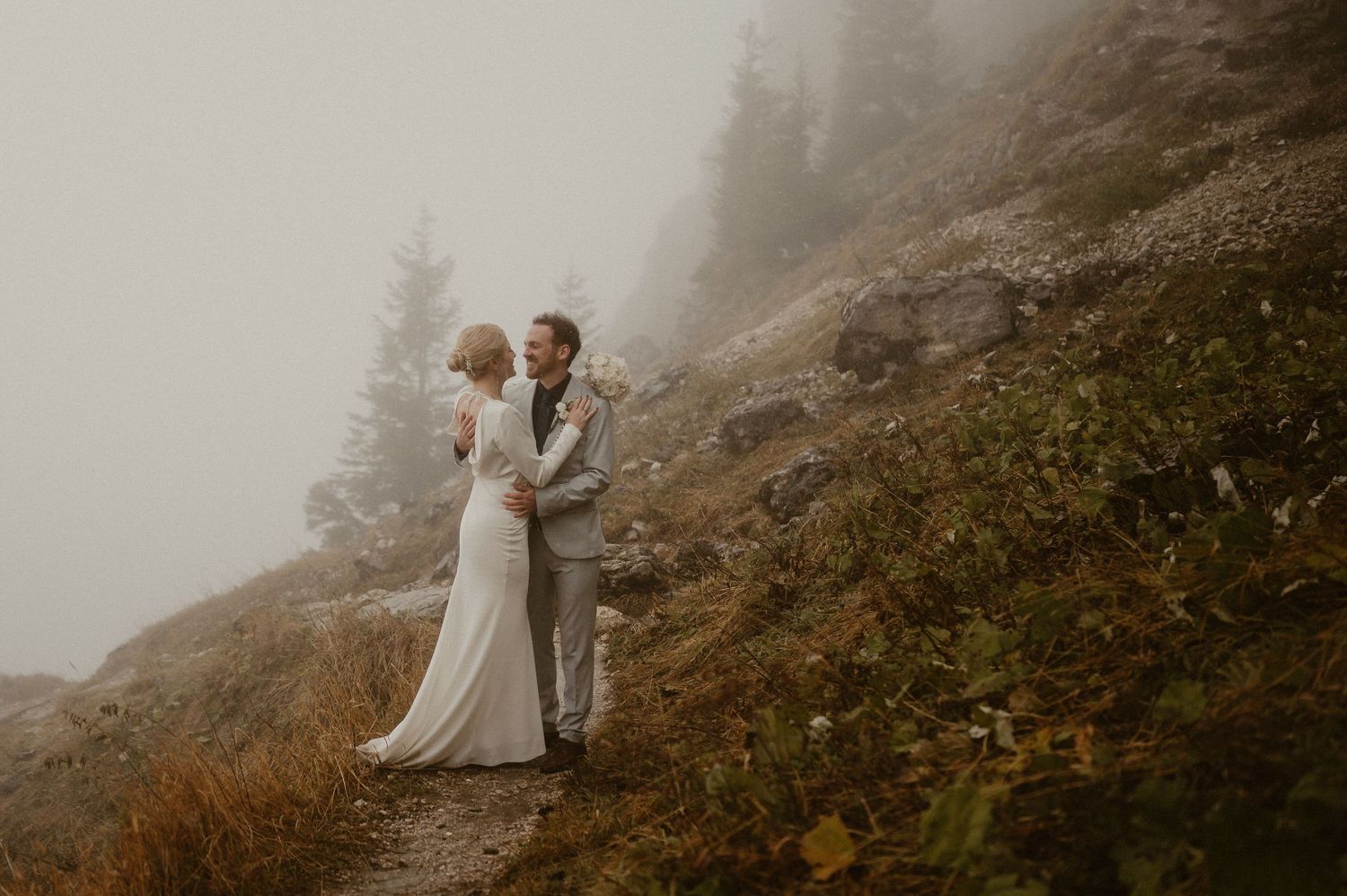 Couple in wedding attire standing together on misty mountain trail.