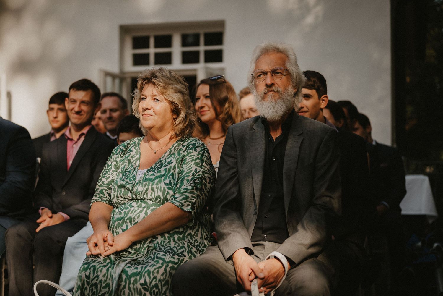 Wedding guests seated in white chairs watching ceremony at outdoor venue.