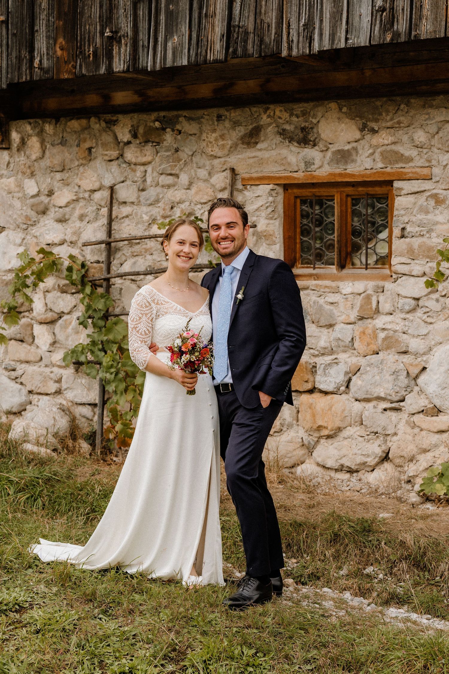 Wedding couple posing against stone wall and wooden barn in outdoor setting.