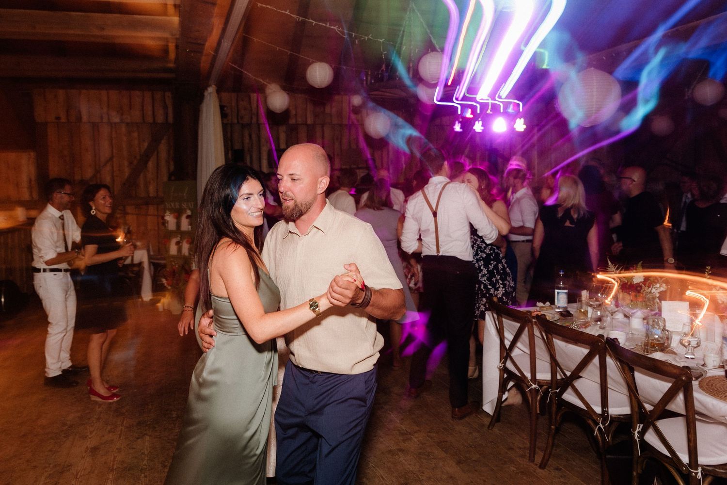 Wedding reception dance party with colorful purple lighting and guests dancing in a rustic venue.