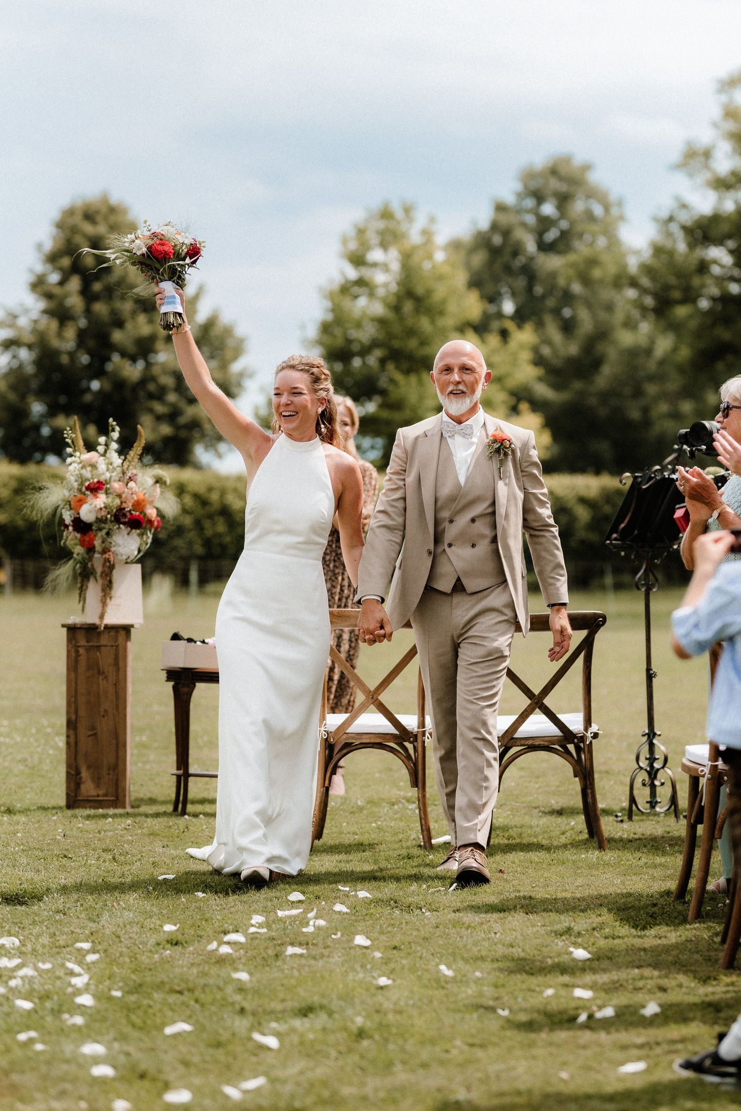 Couple walking down grassy aisle with wooden decor and floral arrangements at outdoor ceremony.