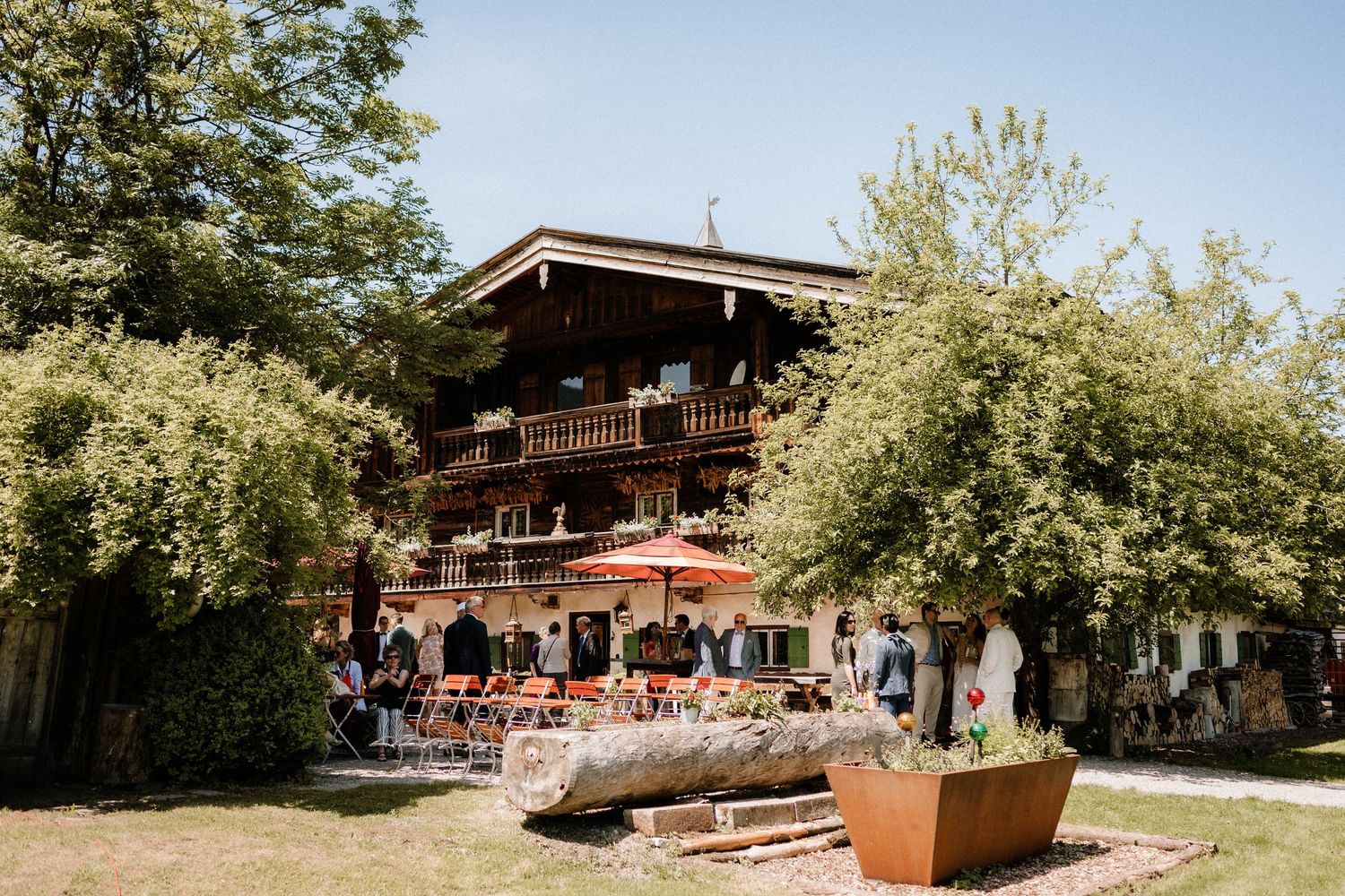Rustic wooden lodge with outdoor seating area surrounded by lush green trees on a sunny day.