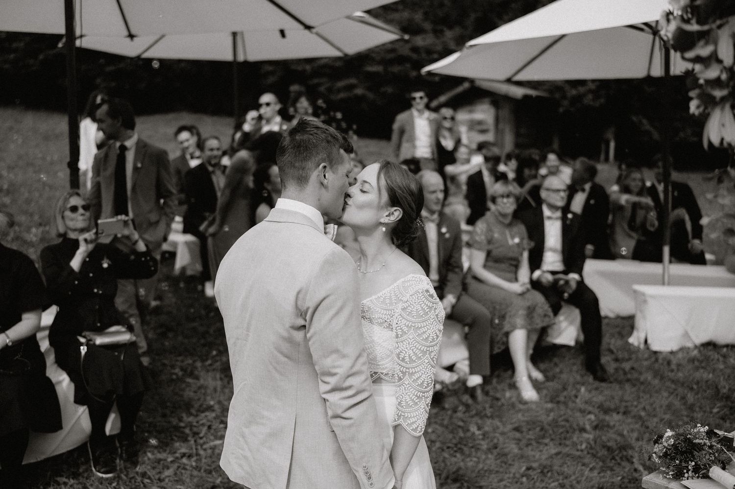 Wedding guests gather outdoors under umbrellas during a celebration in a rustic setting.