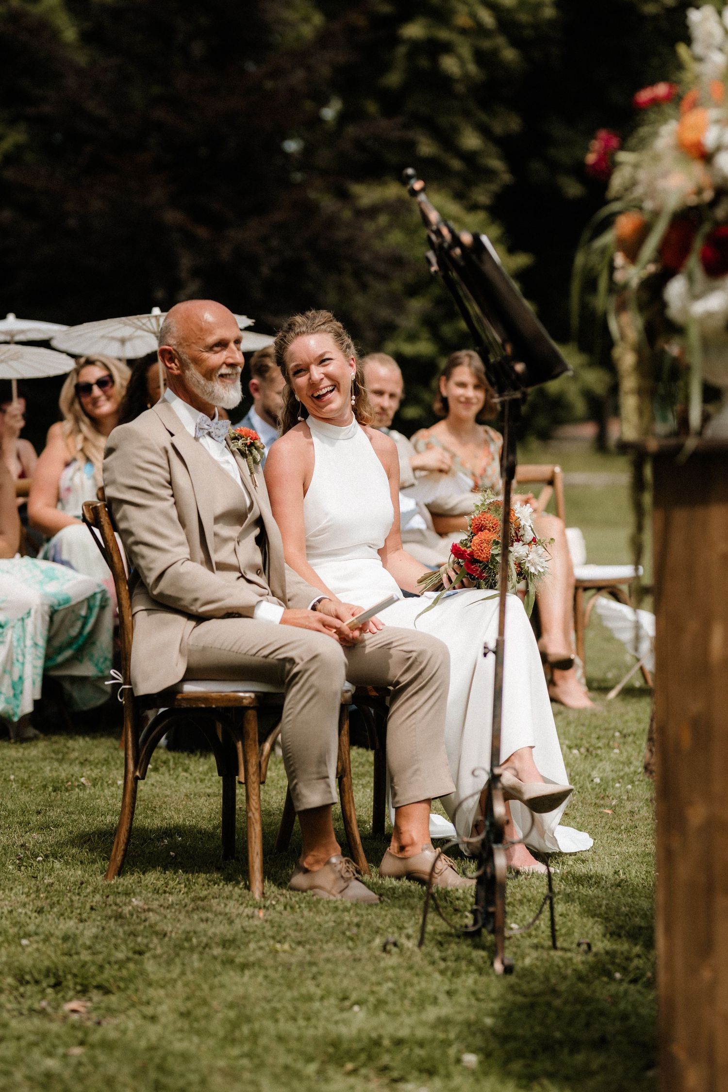 Guests seated on wooden chairs watch an outdoor wedding ceremony on a grassy lawn.