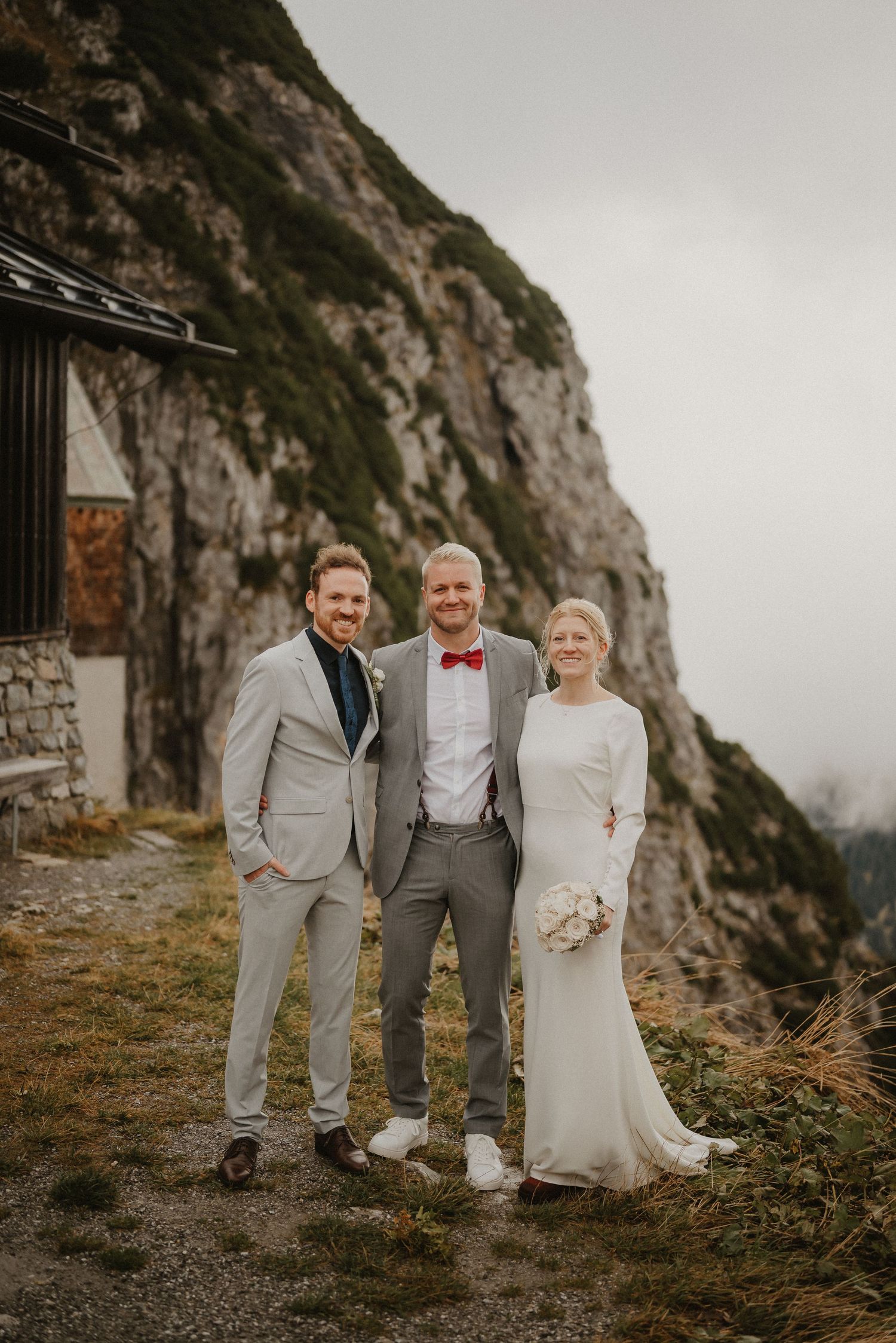 A wedding party poses against a dramatic mountain backdrop near a rustic stone building.