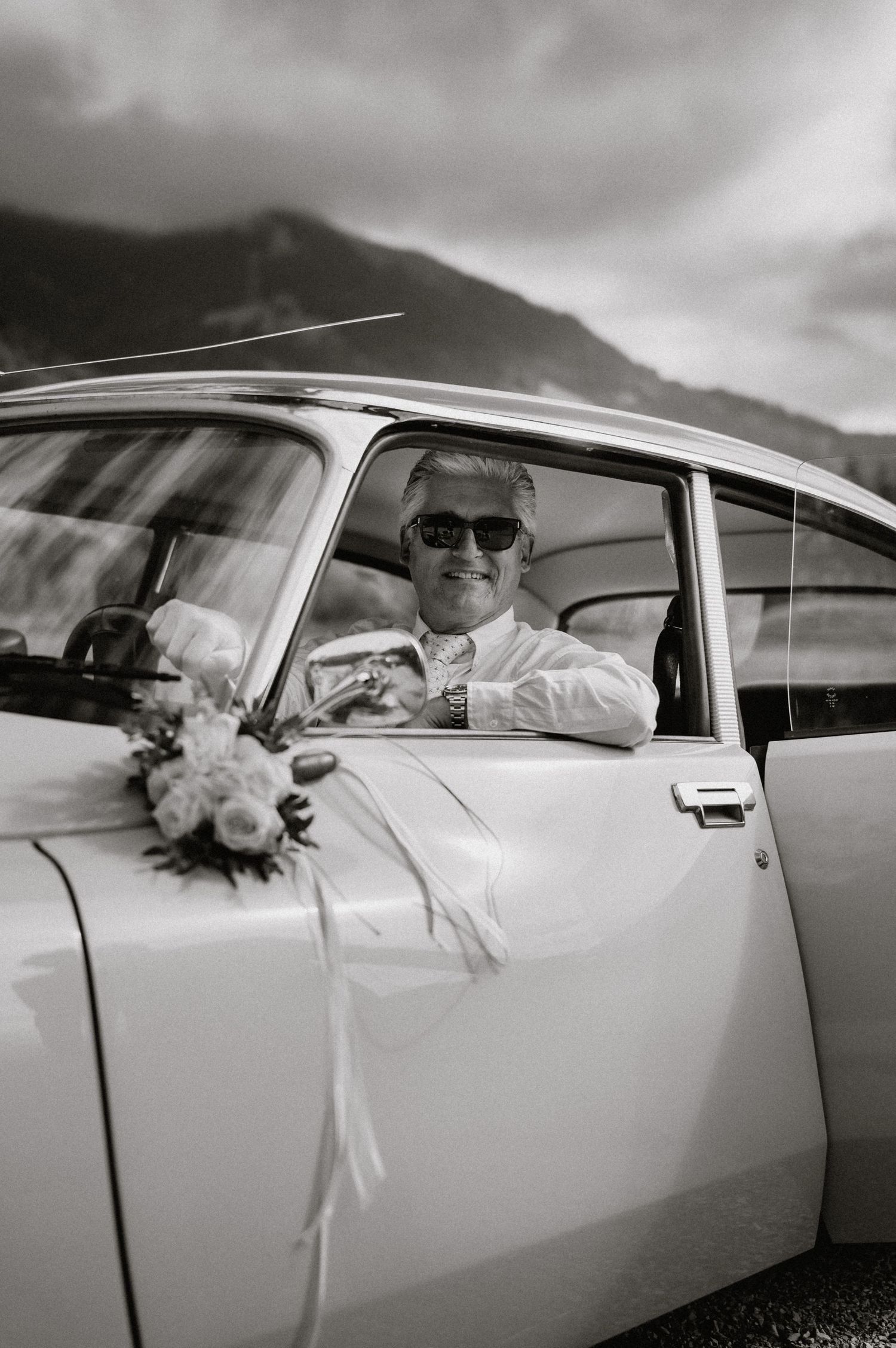 A vintage white car with wedding couple seen through the passenger windows against mountain backdrop.