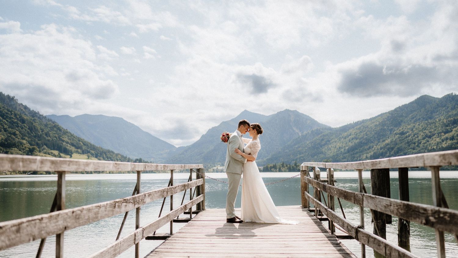 A wedding couple shares an intimate moment on a wooden pier with mountains and lake views.