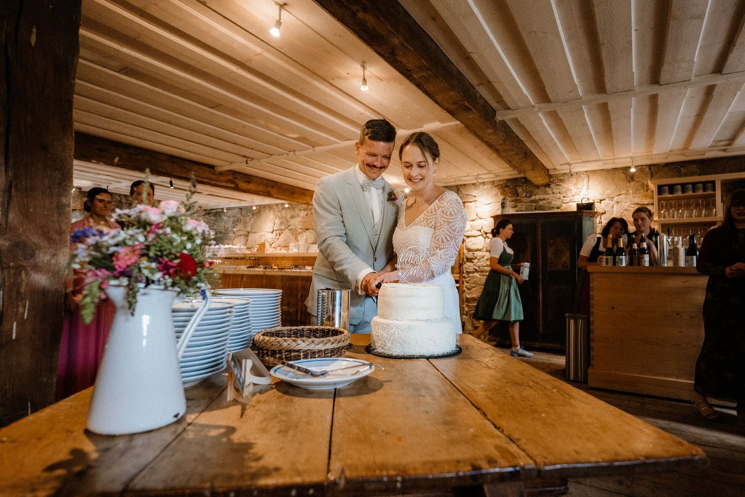 Couple cutting their elegant white wedding cake together in a rustic indoor venue with wooden beams and stone walls.