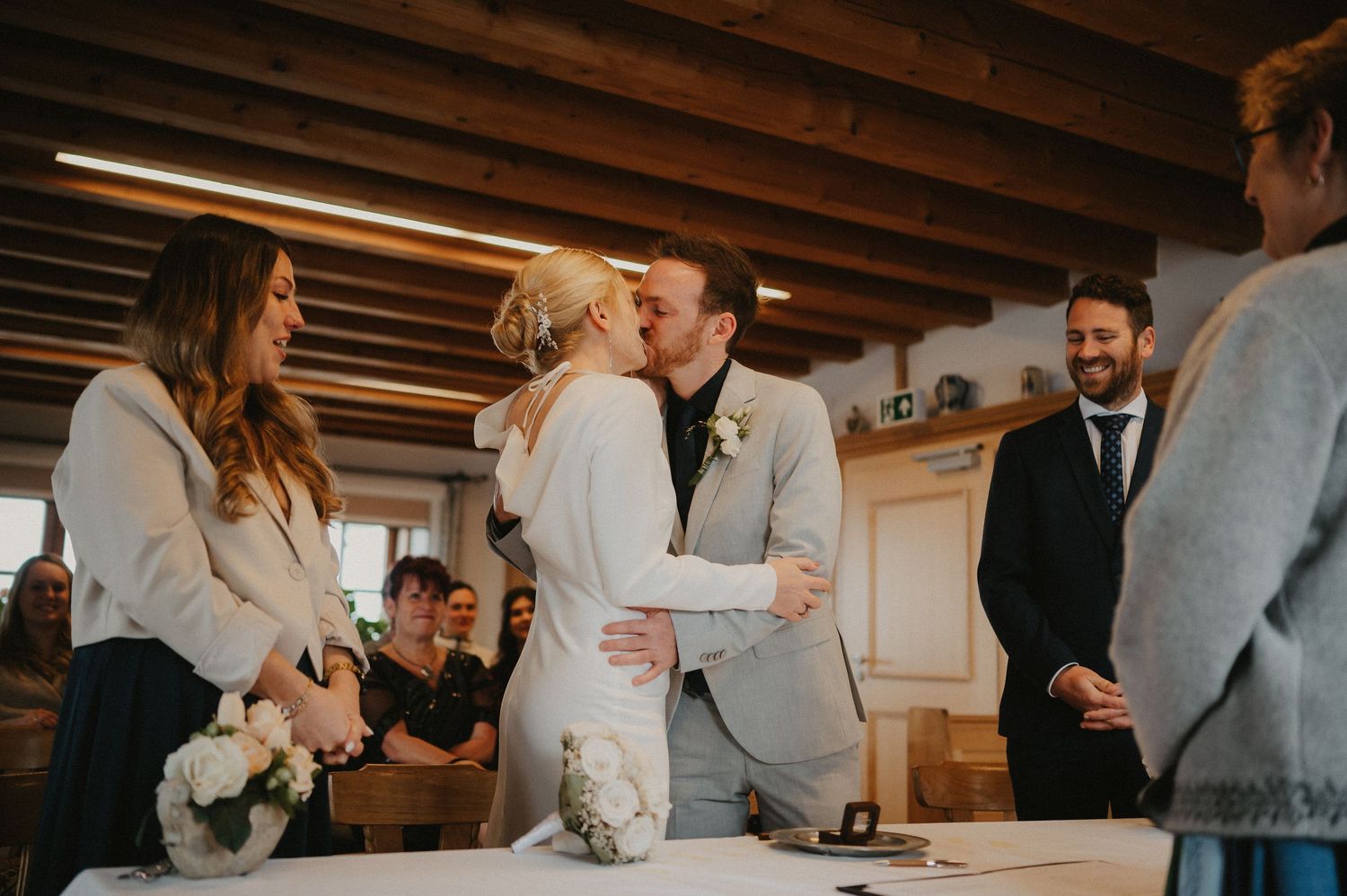 Wedding celebration in a rustic venue with exposed beams as guests gather around a ceremony table.
