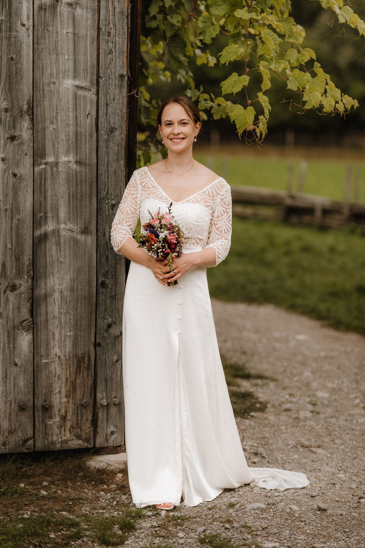 Bride in flowing white dress with lace details stands on gravel path by wooden structure.